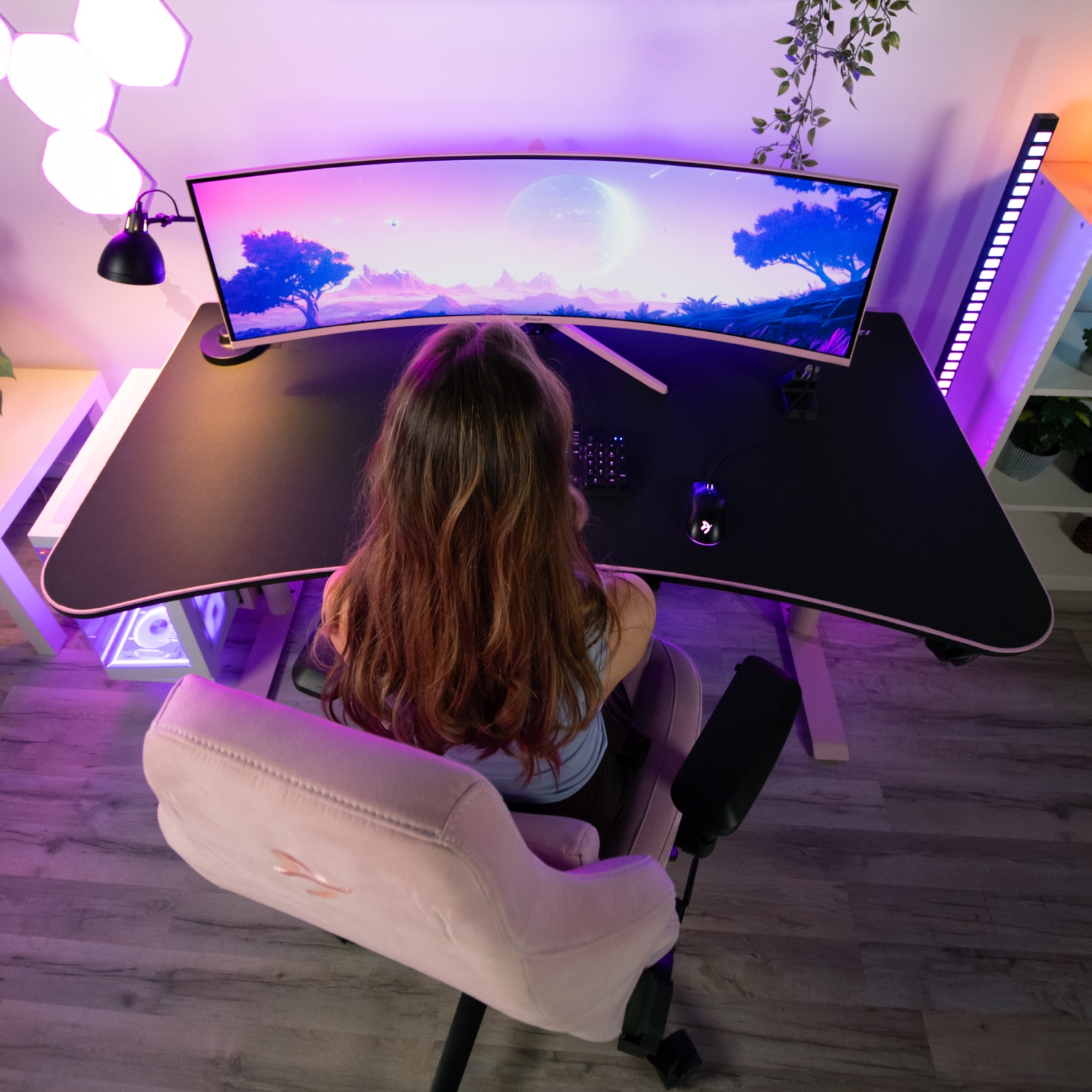 Kobieta siedzi przy biurku do gier z zakrzywionym monitorem. Różowe krzesło, czarne biurko i oświetlenie otoczenia.