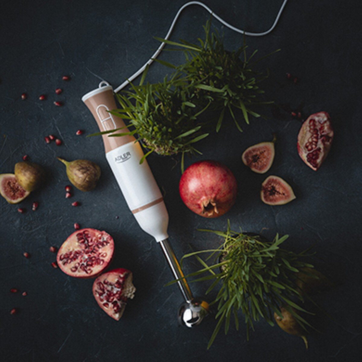 Blender, granaty, figi i zielona trawa na ciemnej powierzchni. Blender jest biało-beżowy. Owoce są przekrojone.