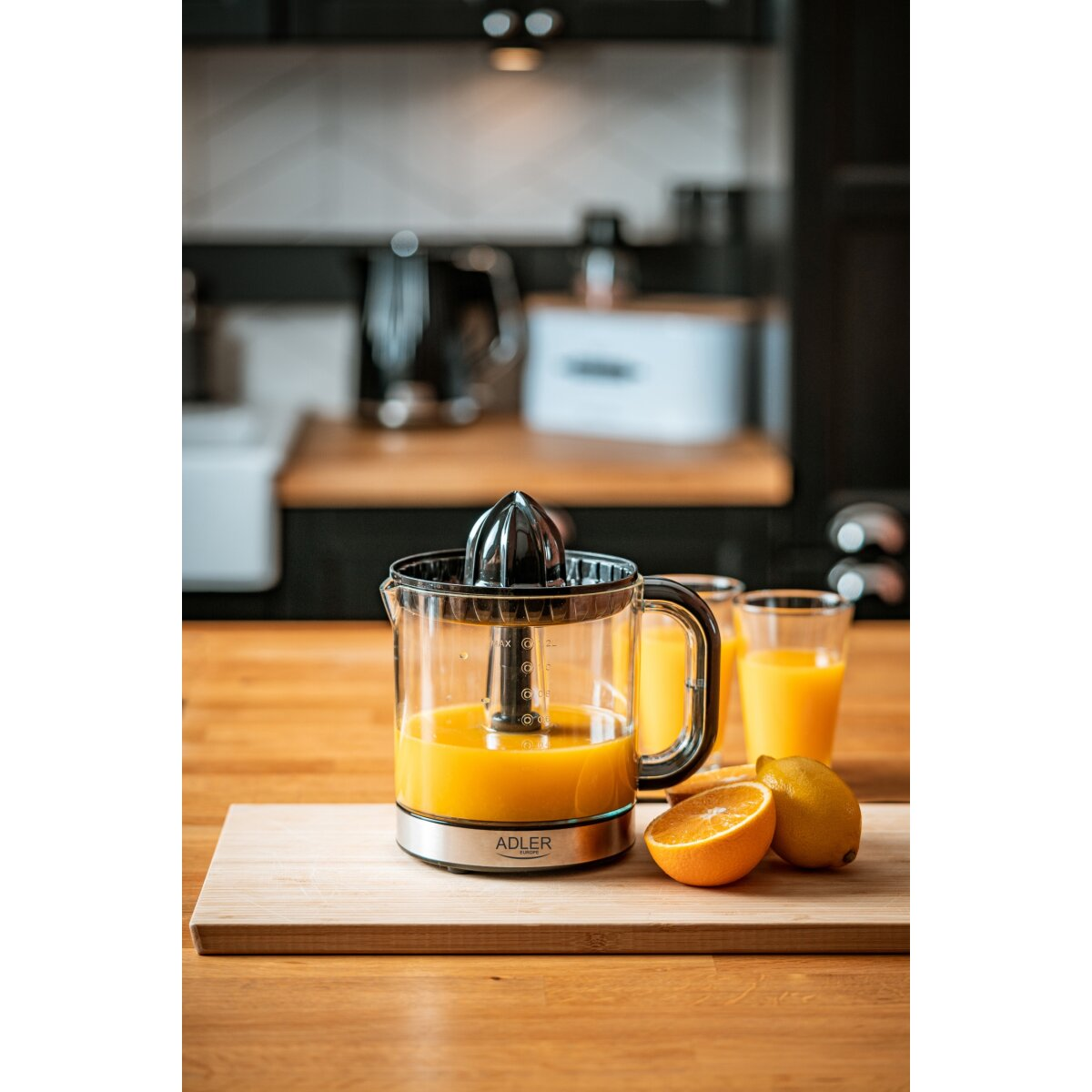 Una exprimidora con zumo de naranja, media naranja, limón sobre una tabla de madera. Dos vasos de zumo al fondo, una cocina.