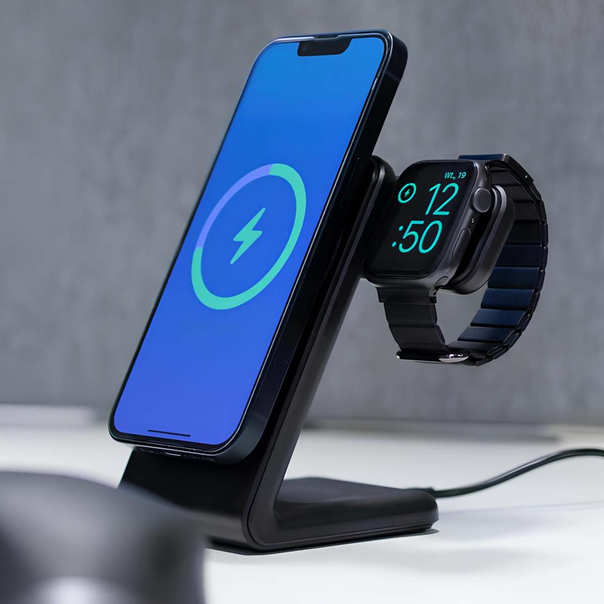 Supporto di ricarica nero con telefono e orologio. Il telefono mostra il simbolo di ricarica, l'orologio mostra l'ora. Tavolo bianco, sfondo grigio.