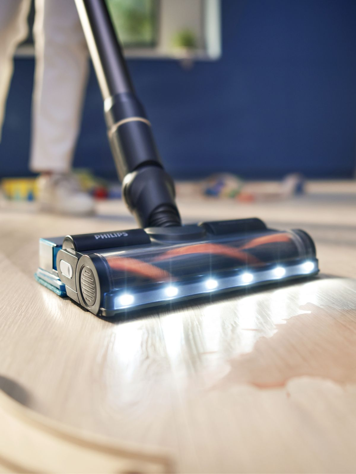 Odkurzacz Philips czyści drewnianą podłogę. Odkurzacz ma światła z przodu. Z tyłu widać osobę w białych spodniach.