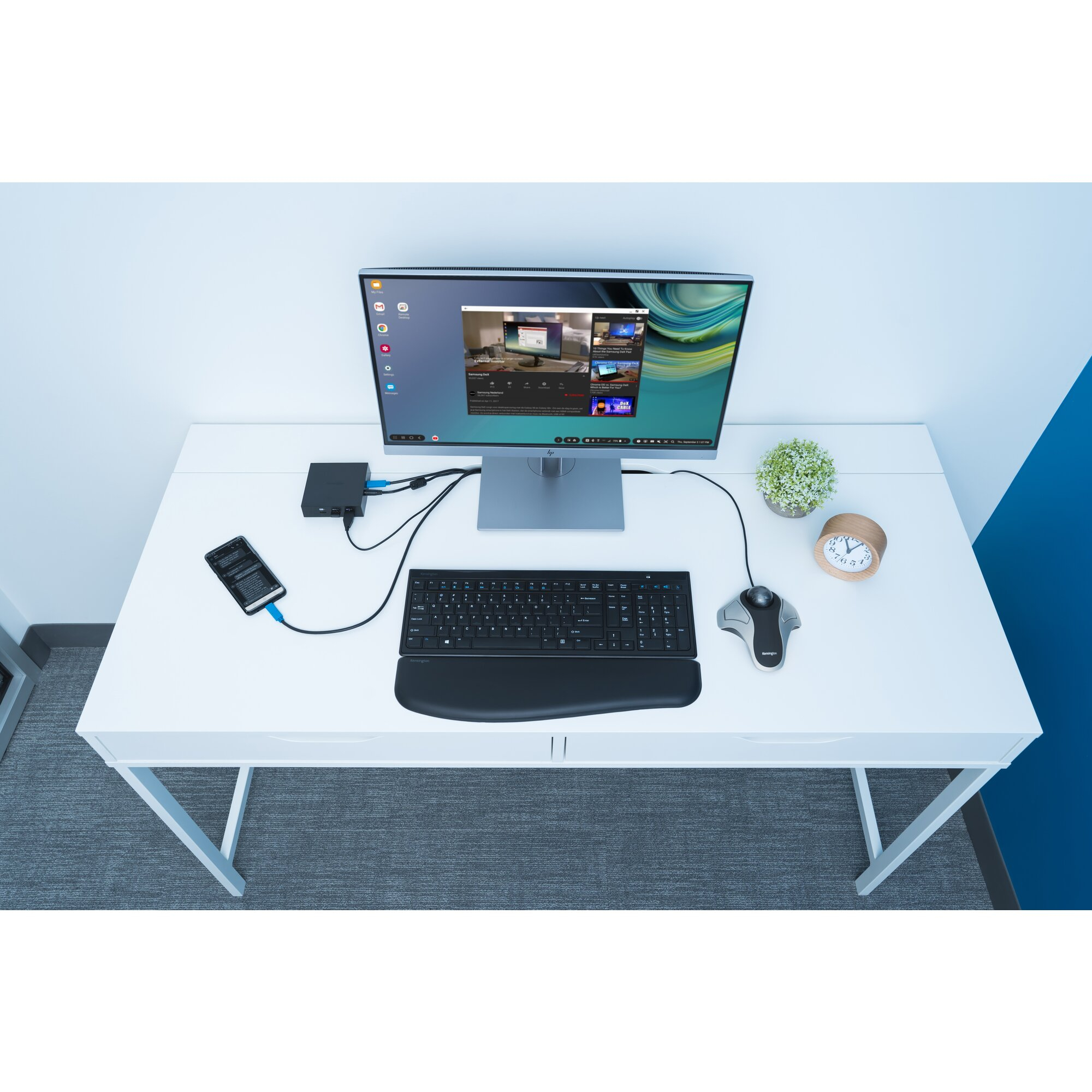 Desk setup with monitor, keyboard, mouse, phone, and clock. Video playing on the screen. White desk and wall.