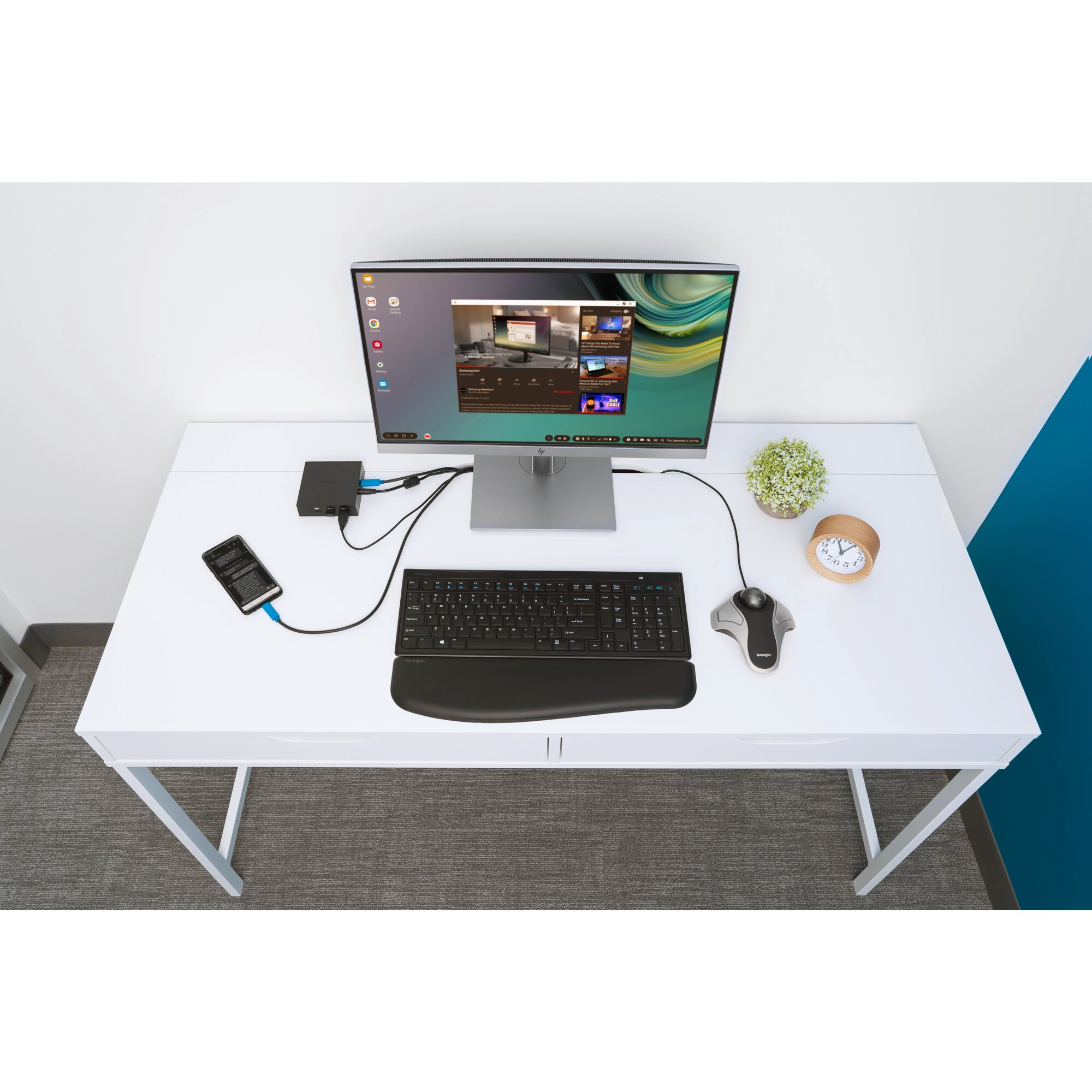 A white desk with a monitor, keyboard, mouse, phone, clock, and a plant. The monitor is displaying a webpage.