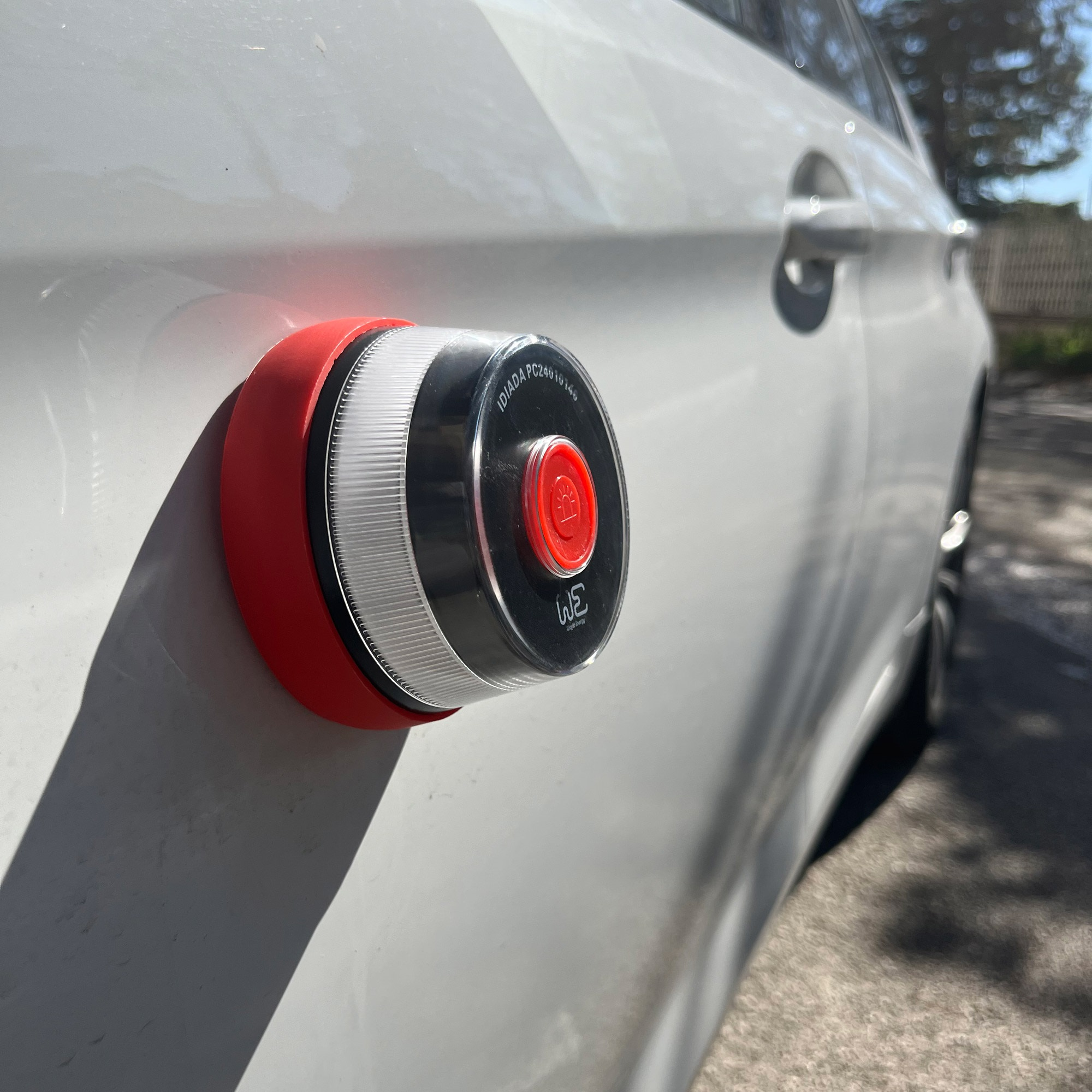 Una luz roja y negra está montada en un coche blanco. La luz tiene una lente transparente y un botón rojo.