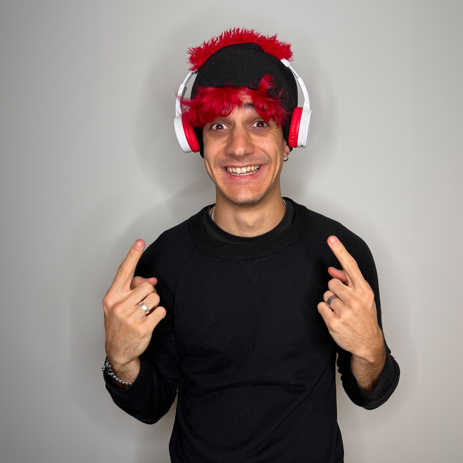 Un uomo sorride, indicando se stesso. Indossa un cappello nero con capelli rossi, cuffie e un maglione nero. Lo sfondo è una parete grigia.