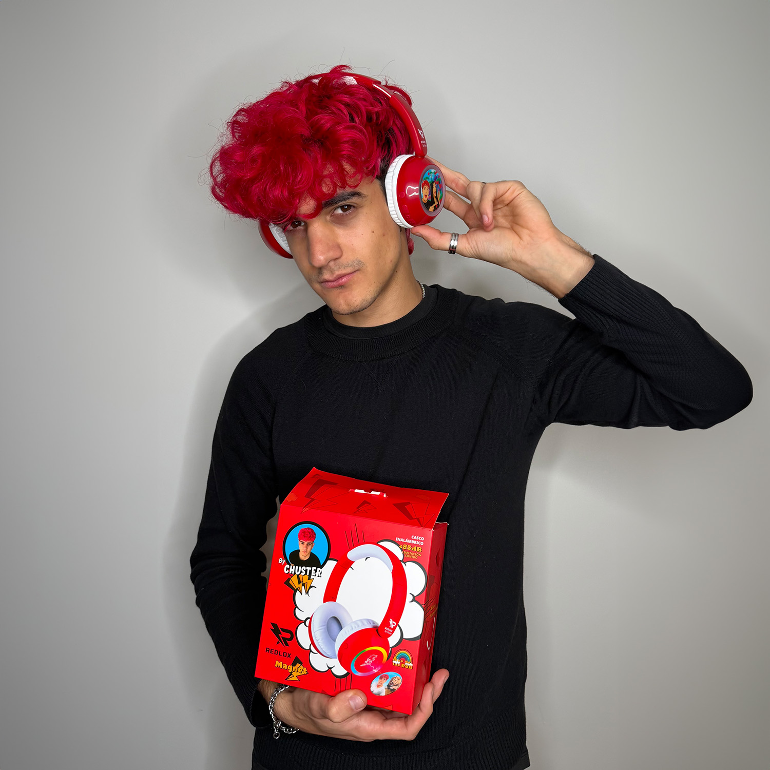 Un uomo con i capelli rossi indossa cuffie rosse e tiene una scatola rossa con le cuffie, in piedi davanti a un muro bianco.