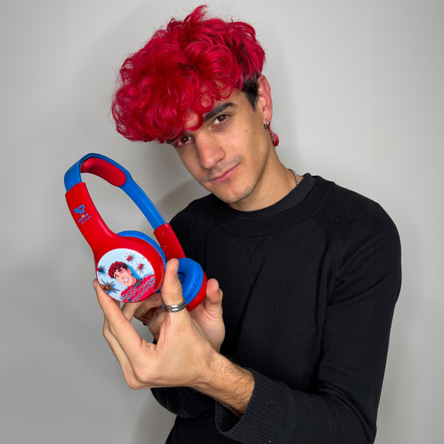 Un hombre con pelo rojo sostiene auriculares rojo/azules. Lleva un suéter negro y joyas plateadas. El fondo es una pared lisa.