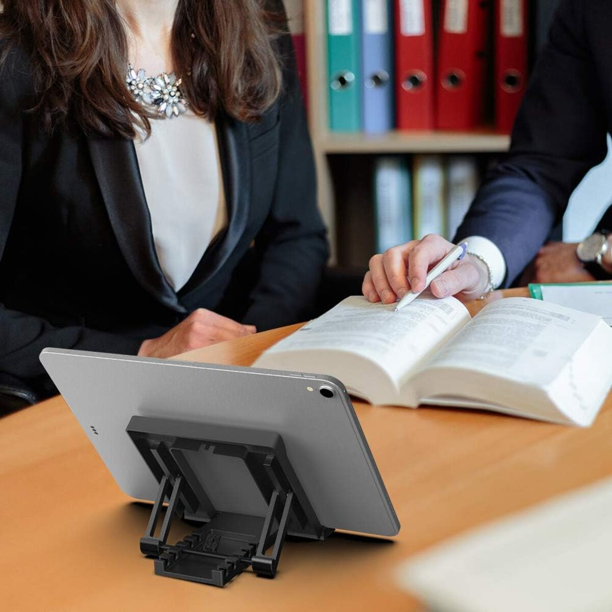 Ein Tablet auf einem Ständer, mit zwei Personen in Anzügen, von denen eine in ein Buch schreibt. Bücherregale im Hintergrund.