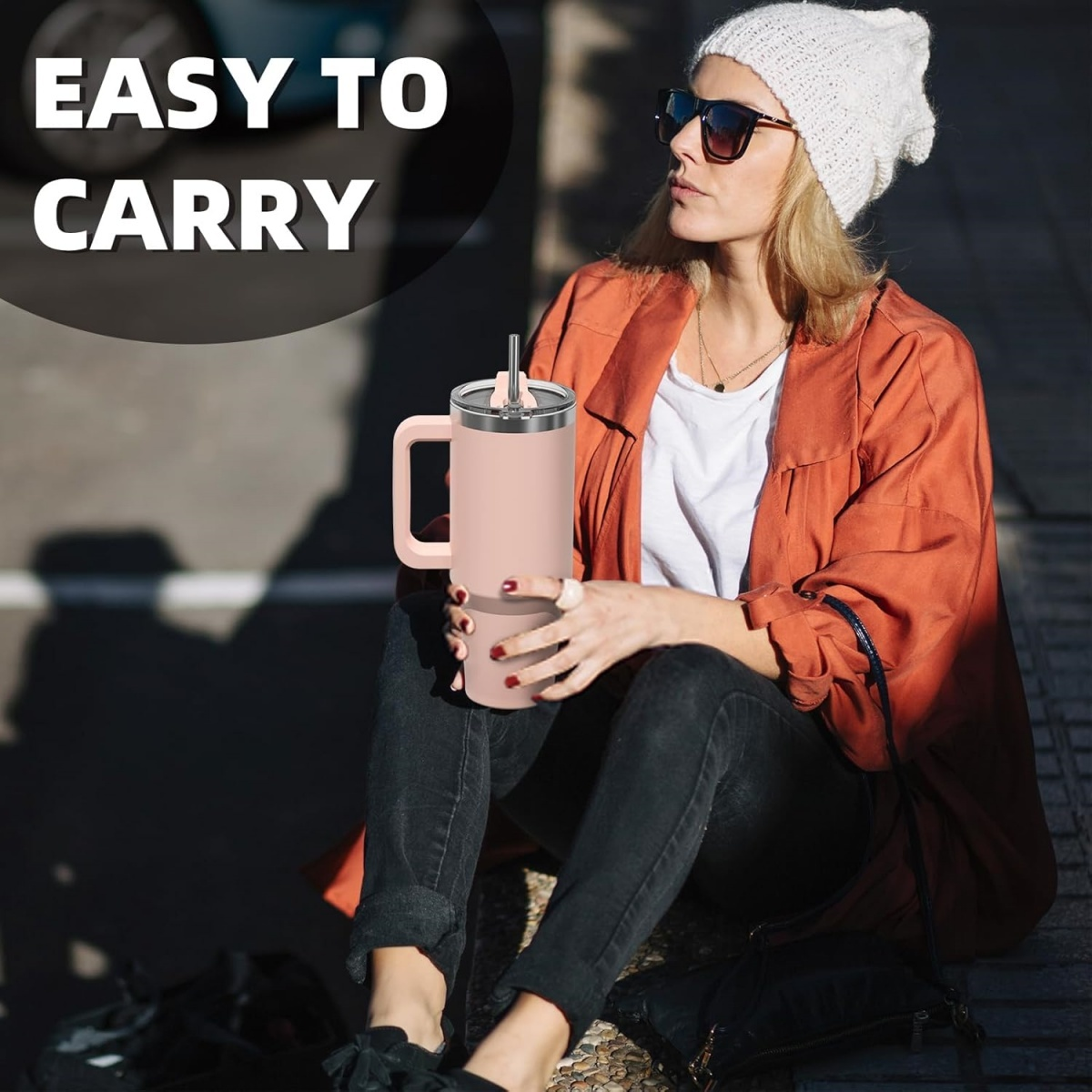 Eine Frau mit Sonnenbrille und Mütze hält einen rosa Becher mit Henkel. Sie trägt eine rote Jacke und schwarze Jeans.