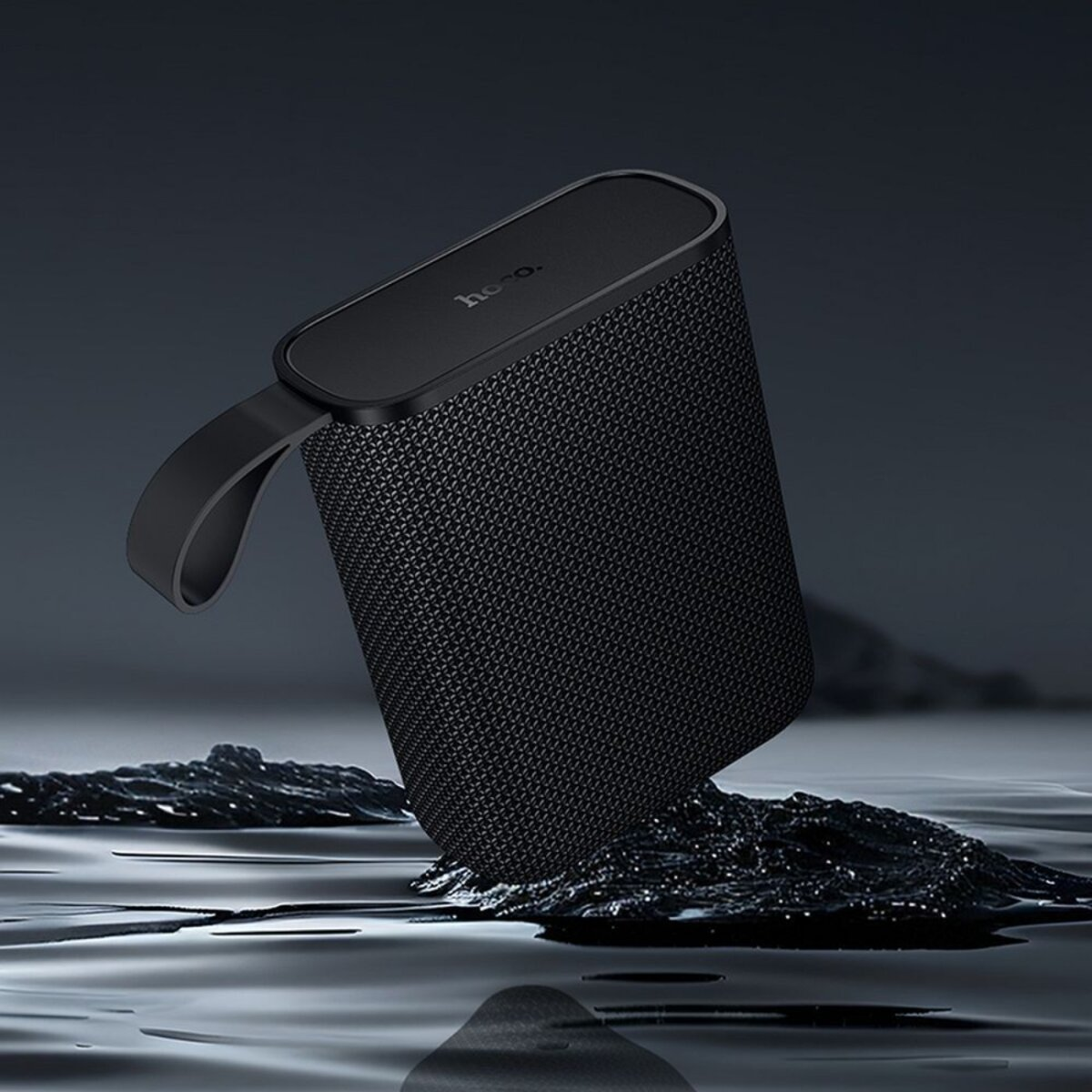 Un altoparlante nero con manico poggia su una superficie scura, riflettendosi nell'acqua. L'altoparlante ha una trama a rete e il logo "hoco."
