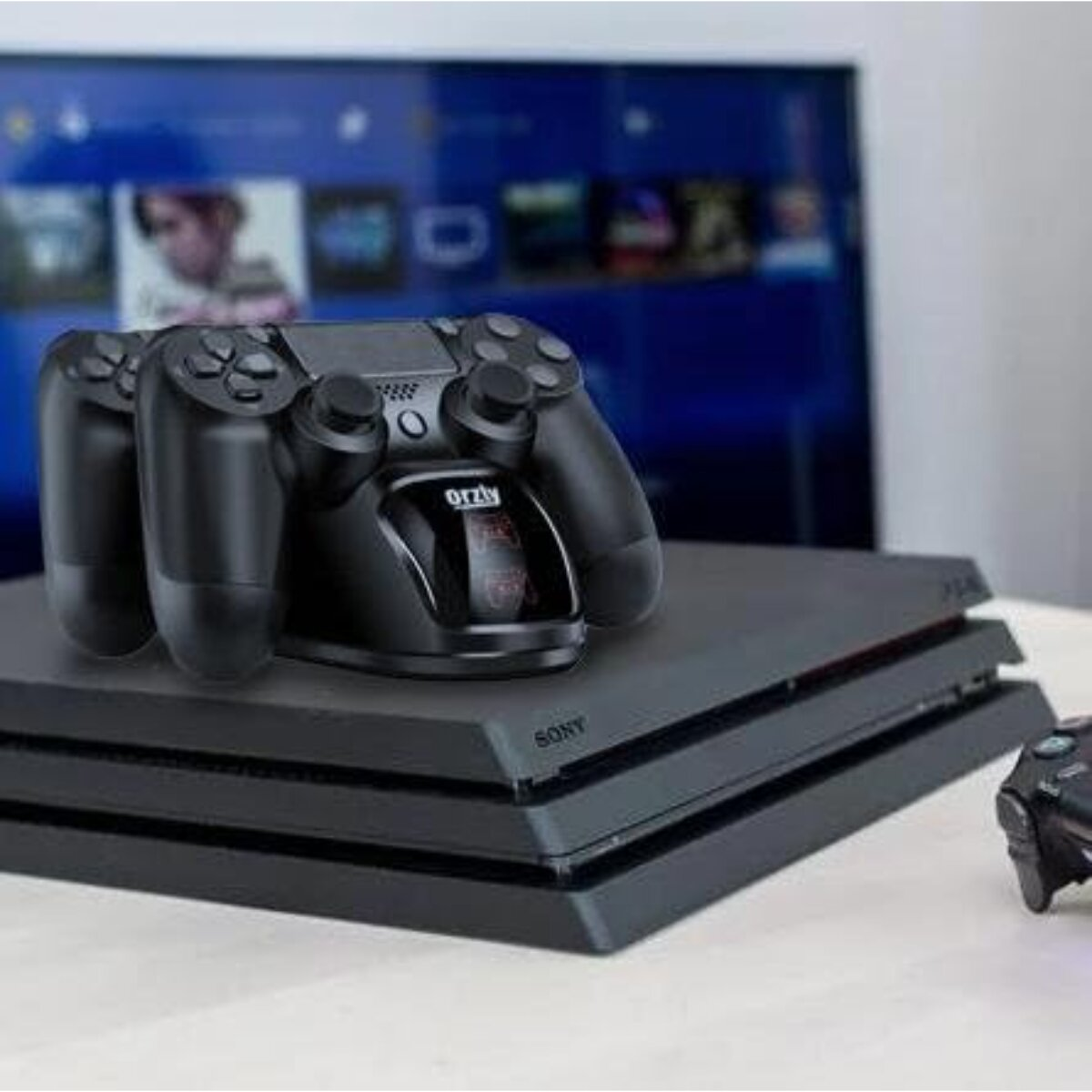 Eine schwarze PlayStation-Konsole mit zwei Controllern, die oben aufgeladen werden. Im Hintergrund ein Fernseher mit einer Spieloberfläche.