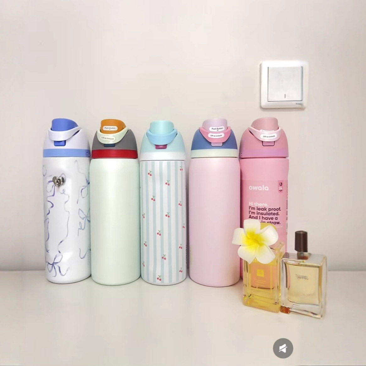 Fünf bunte Wasserflaschen, zwei Parfümflaschen und eine Blume stehen auf einer weißen Oberfläche. An der weißen Wand befindet sich ein Lichtschalter.