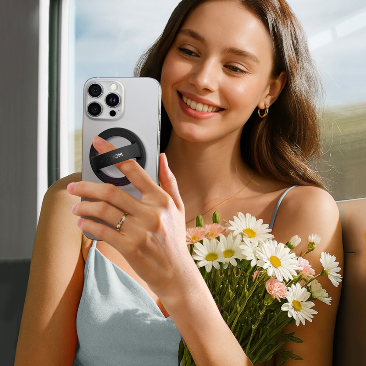 Frau hält Telefon mit Ringhalter, lächelt und Blumen. Ring am Finger. Im Hintergrund ein Fenster.