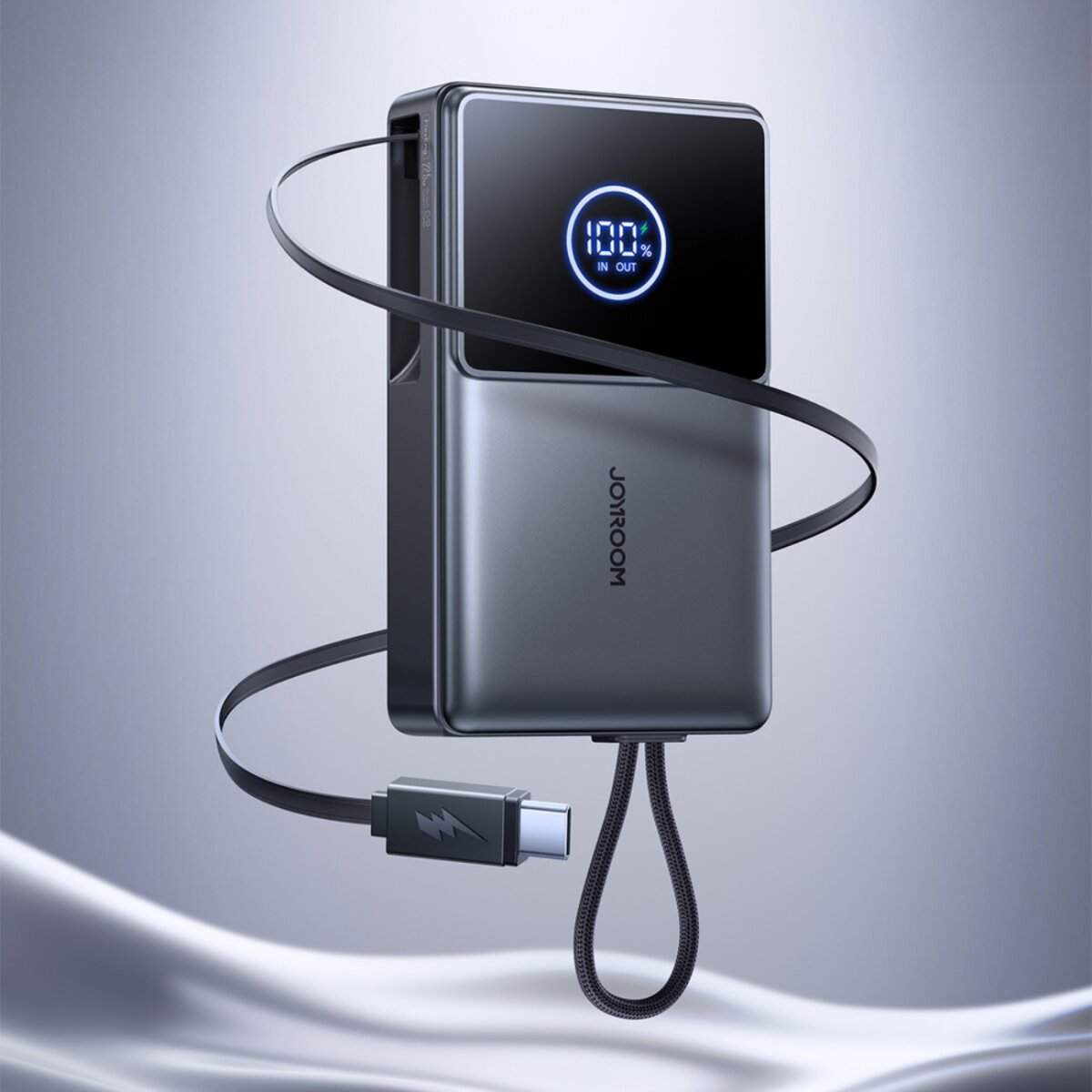 Un power bank grigio con display digitale che mostra il 100%. Un cavo nero è collegato, con connettore USB-C. Il marchio è Joyroom.