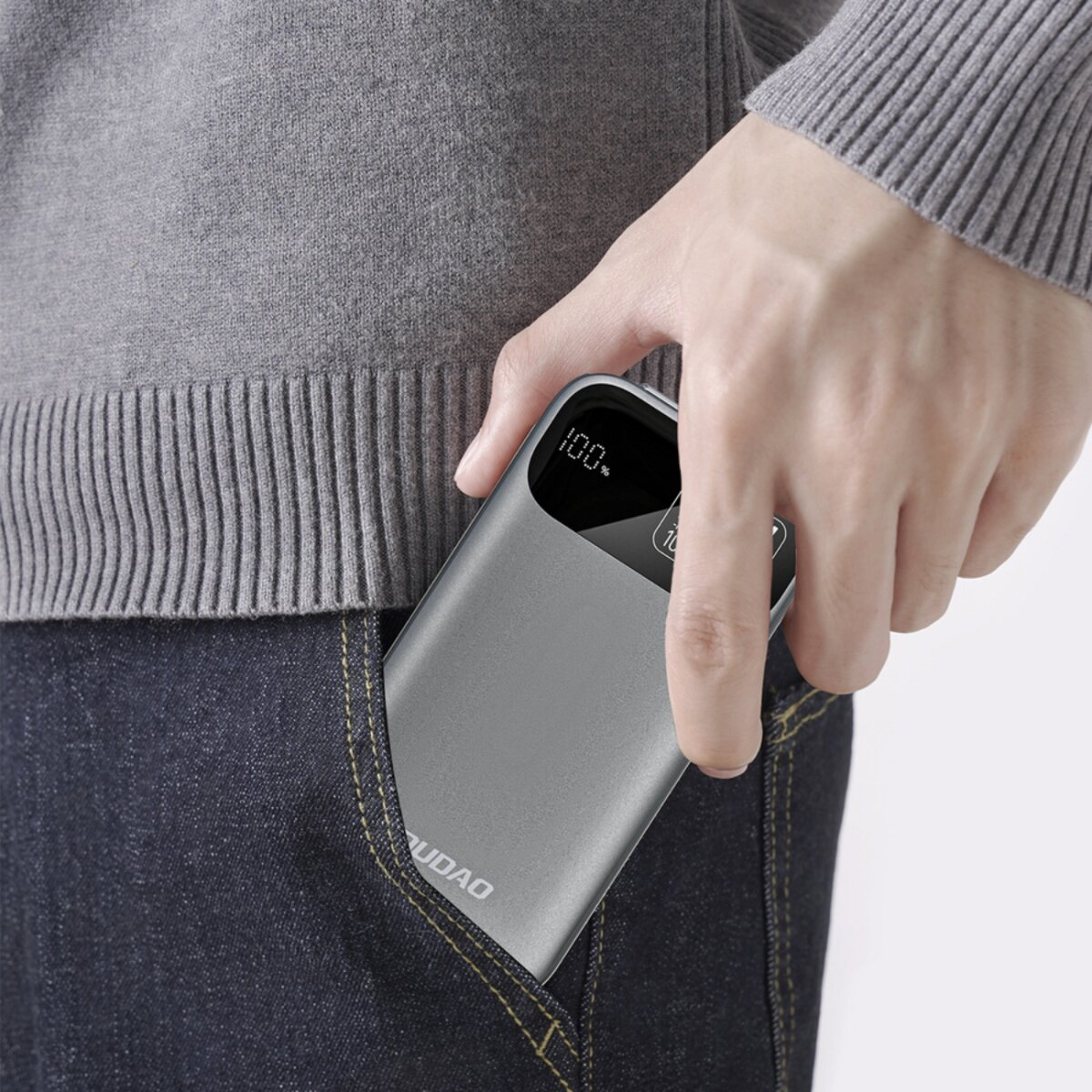 Una persona mette un power bank grigio in una tasca dei jeans. Lo schermo del power bank mostra 100%. La persona indossa un maglione grigio.