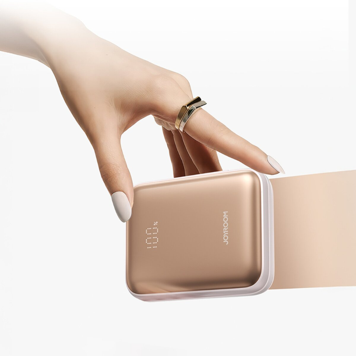 Una mano con un anello tiene un power bank dorato. Il display mostra il 100%. Lo sfondo è bianco e marrone chiaro.