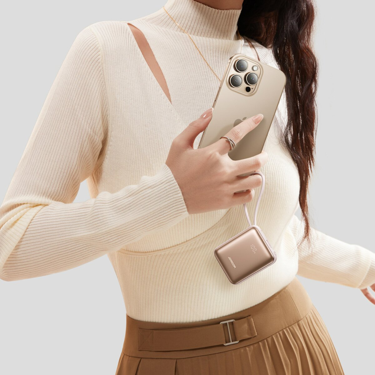 Una donna tiene in mano un telefono d'oro e un power bank. Indossa un maglione color crema e una gonna marrone. Sono visibili anche una collana e un anello.