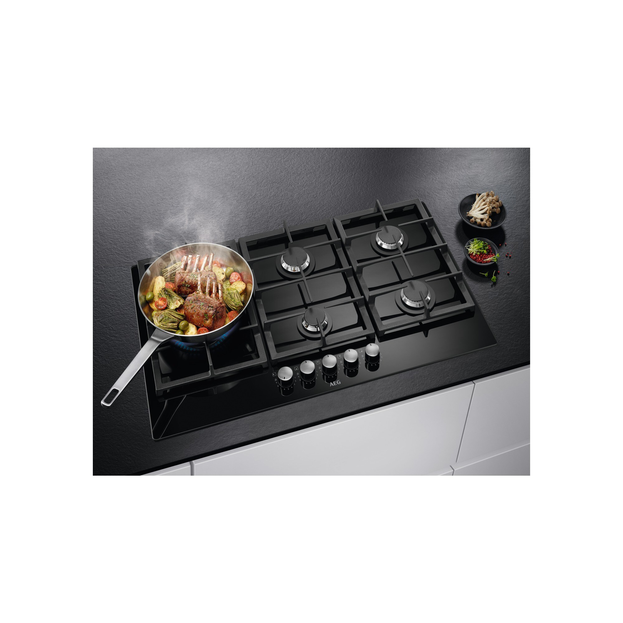 Czarna płyta grzewcza z patelnią z ugotowanym jedzeniem po lewej. Grzyby i zioła po prawej. Białe szafki kuchenne poniżej.