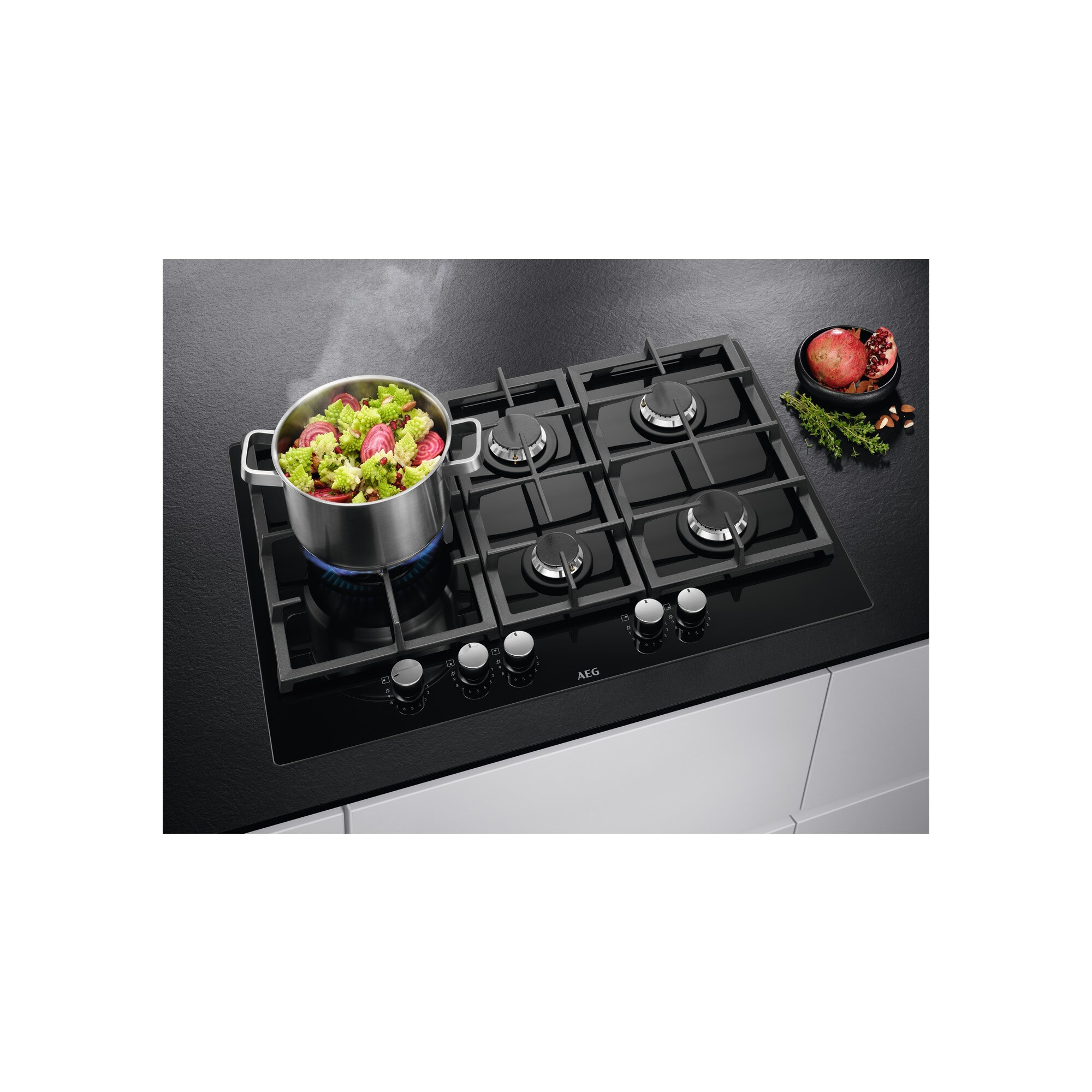 Czarna płyta kuchenna z garnkiem warzyw parujących. Miska z granatem i ziołami obok.