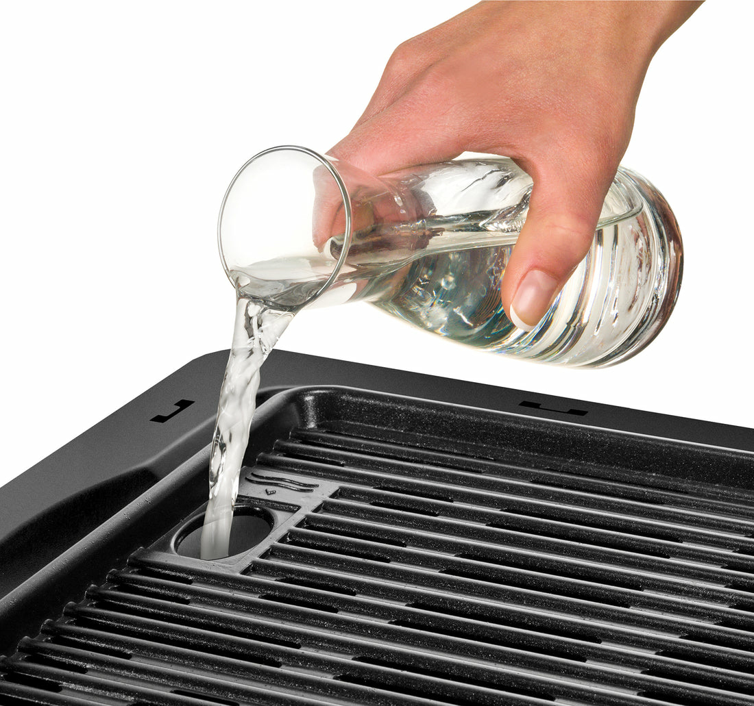 Eine Hand gießt Wasser aus einer Glaskaraffe in einen schwarzen Grill. Wasser strömt herunter. Der Hintergrund ist weiß.