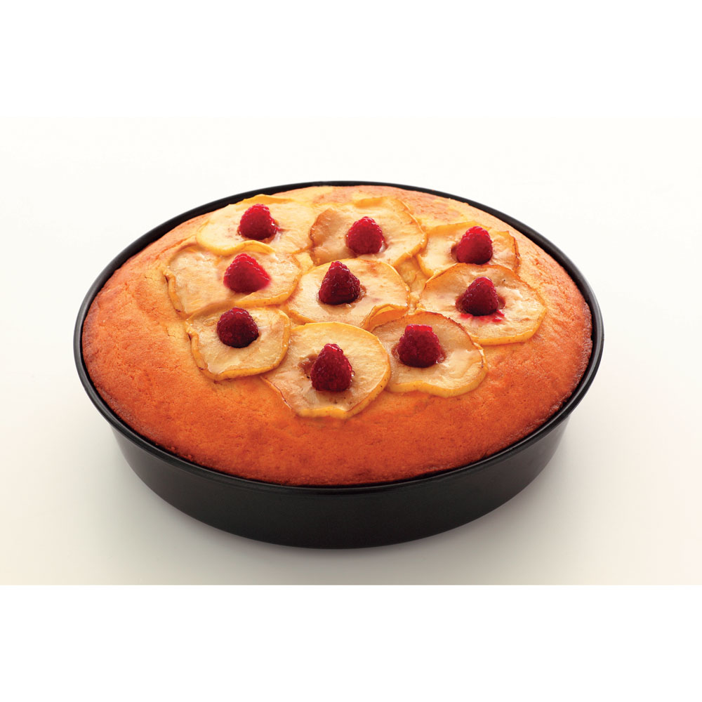 Una torta rotonda in una teglia nera, guarnita con fette di pera e lamponi su una superficie bianca. La torta ha una crosta dorata.