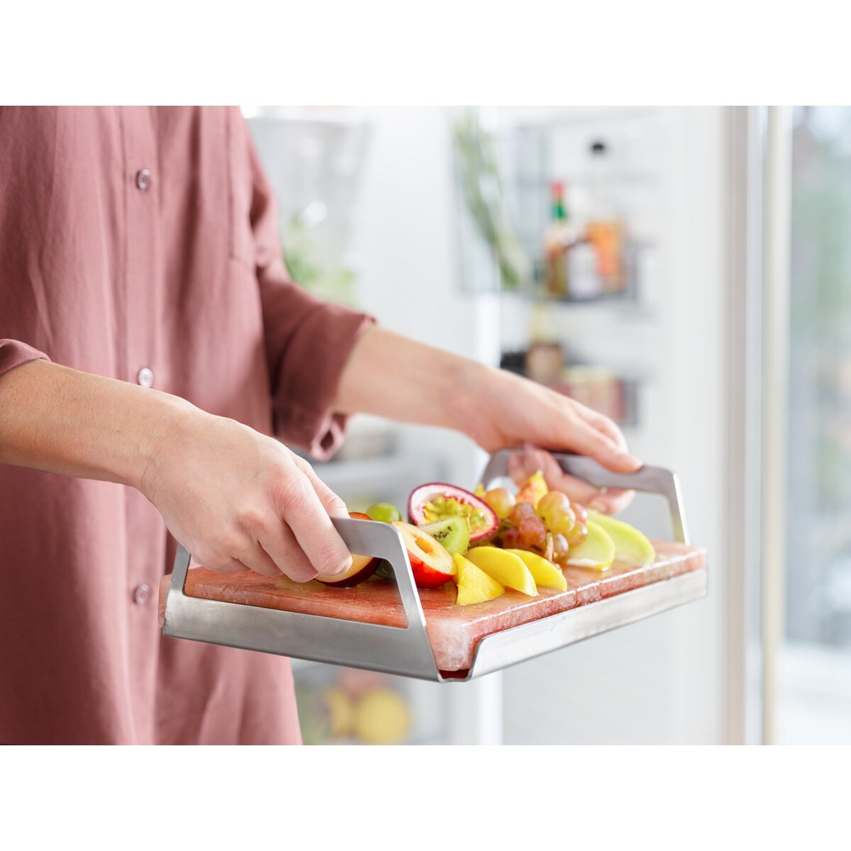 Una persona tiene in mano un vassoio di frutta tagliata: uva, kiwi, frutto della passione, mela e mango. Il vassoio ha manici in metallo. Lo sfondo è un frigorifero.