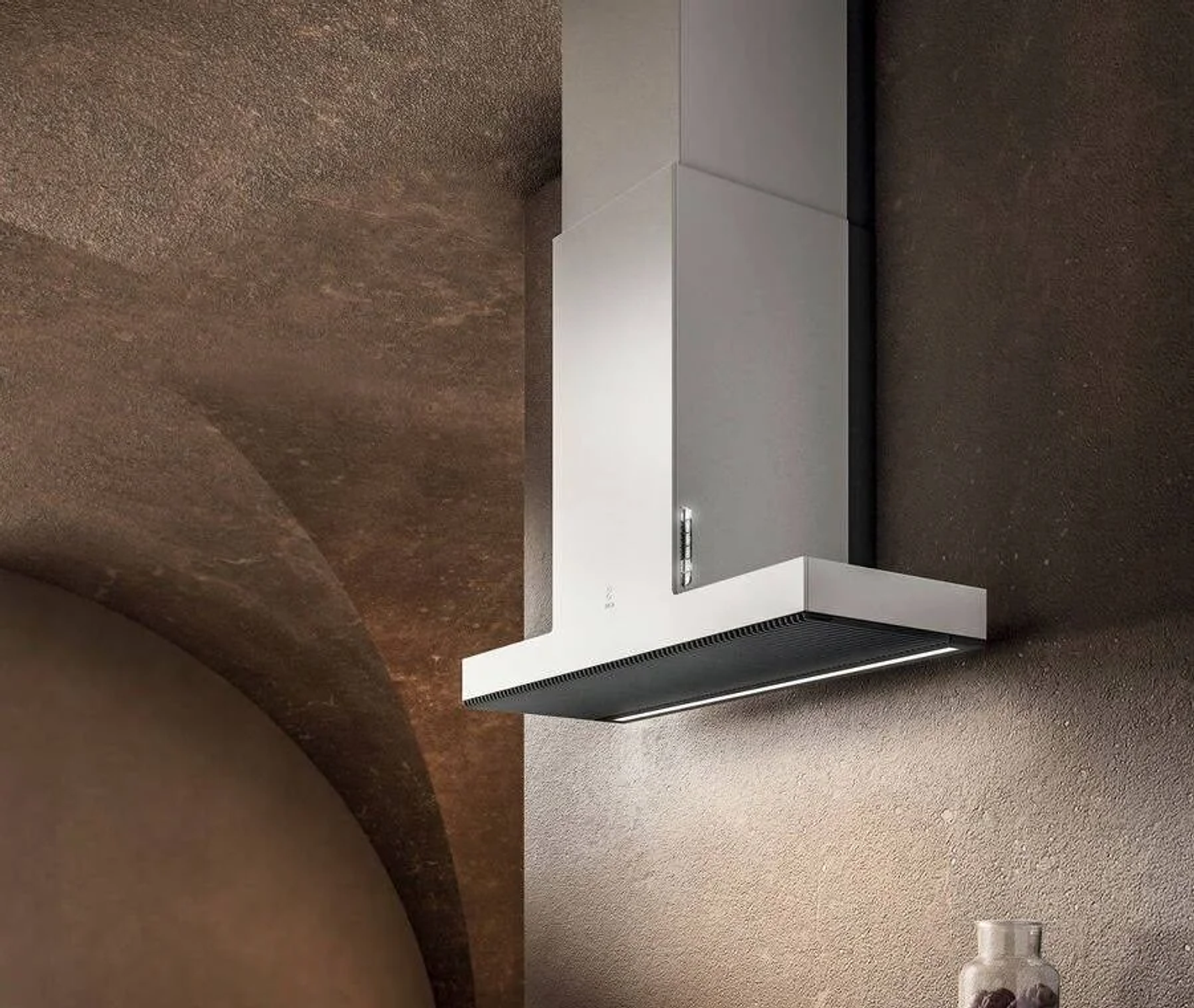 Una moderna cappa da cucina pende dal soffitto. La cappa è bianca. Lo sfondo è una parete e un soffitto marroni strutturati.