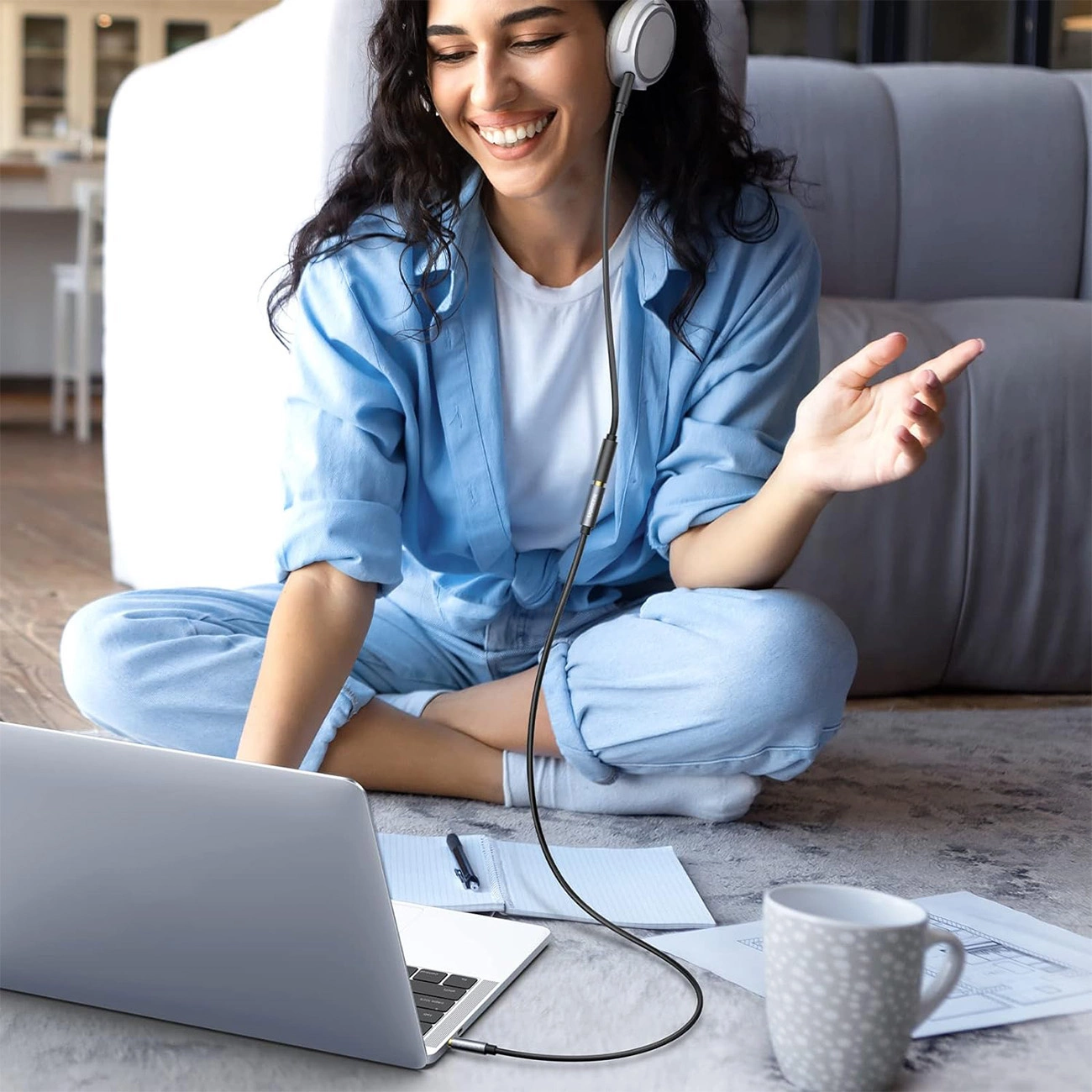Kobieta w słuchawkach, uśmiechnięta, siedzi na podłodze z laptopem, filiżanką kawy i papierami. Ma na sobie niebieskie ubrania.