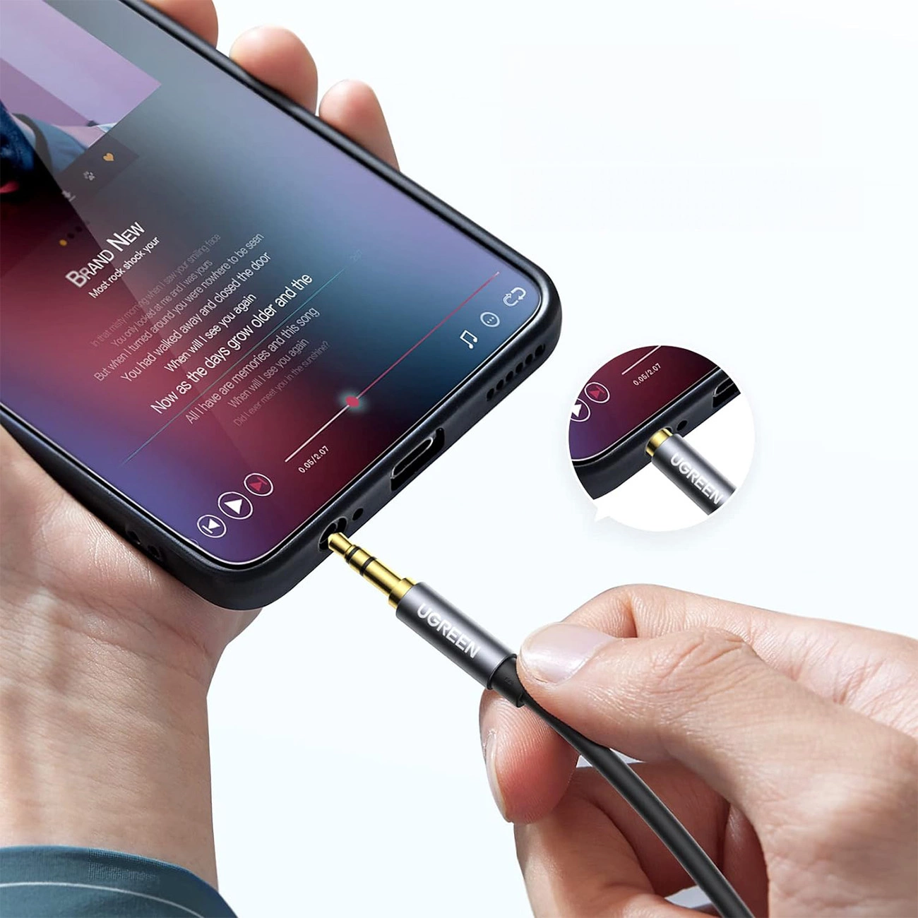 Ręka trzyma telefon odtwarzający muzykę, podłączając kabel audio UGREEN. Telefon wyświetla tekst piosenki. Zbliżenie na kabel.