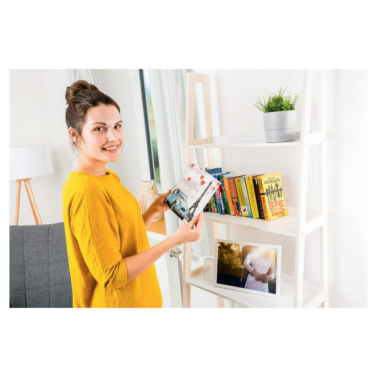 Una mujer de amarillo sostiene un libro con la Torre Eiffel. Estantería con libros y una foto enmarcada en el fondo.