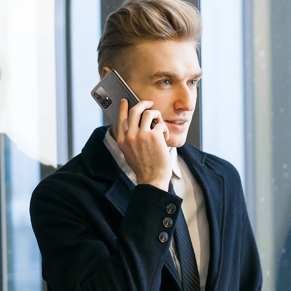 Ein Mann im Anzug telefoniert. Er hat blondes Haar und blaue Augen. Er trägt einen schwarzen Mantel und eine Krawatte. Der Hintergrund ist verschwommen.