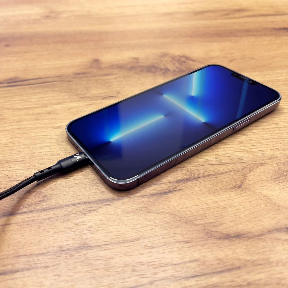 Ein iPhone liegt auf einer Holzoberfläche und ist angeschlossen. Der Bildschirm ist blau mit Lichtreflexen. Ein schwarzes Kabel verbindet sich mit dem Telefon.