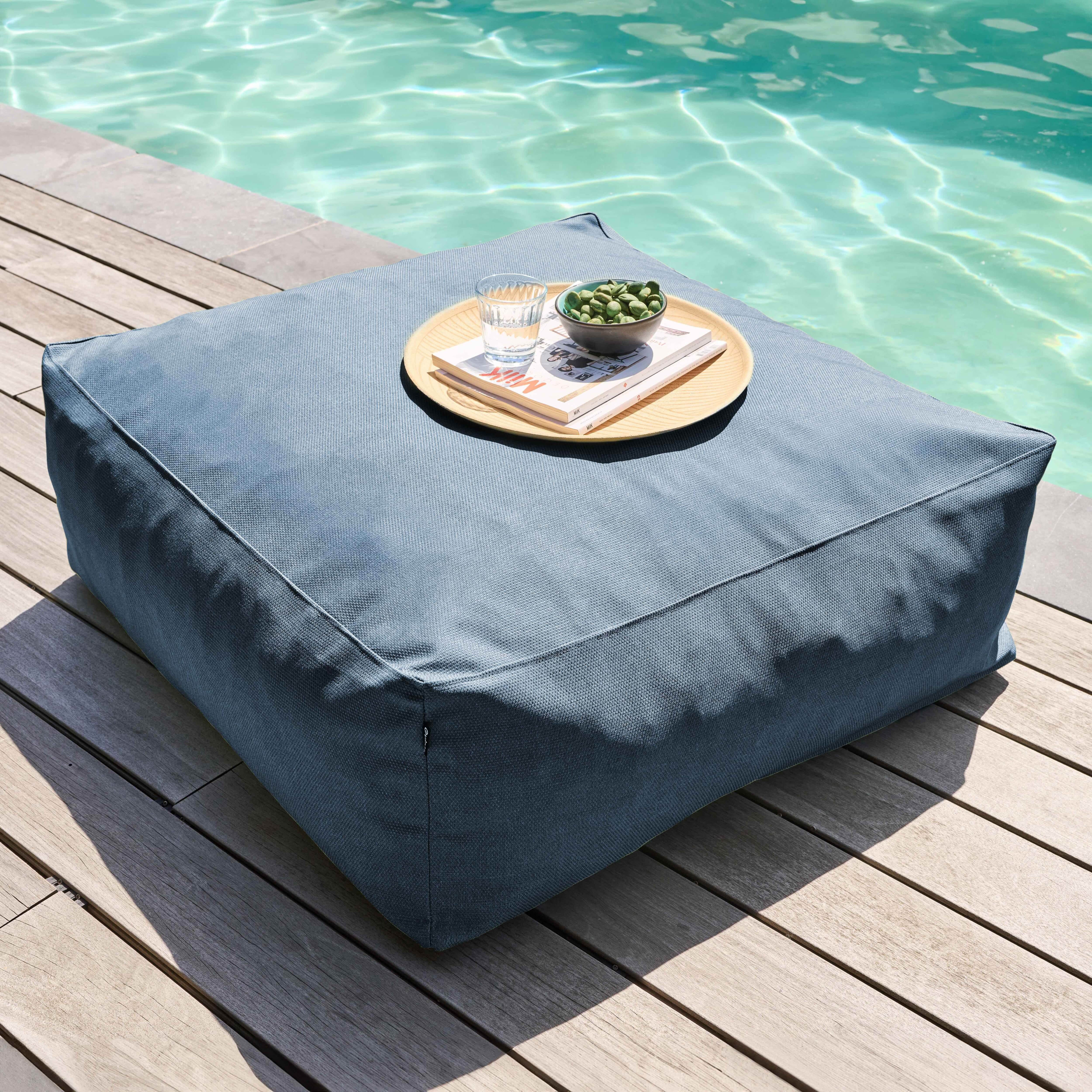 Blaues quadratisches Kissen auf Holzplanken, mit einem Tablett mit Snacks und einem Wasserglas. Poolwasser im Hintergrund.