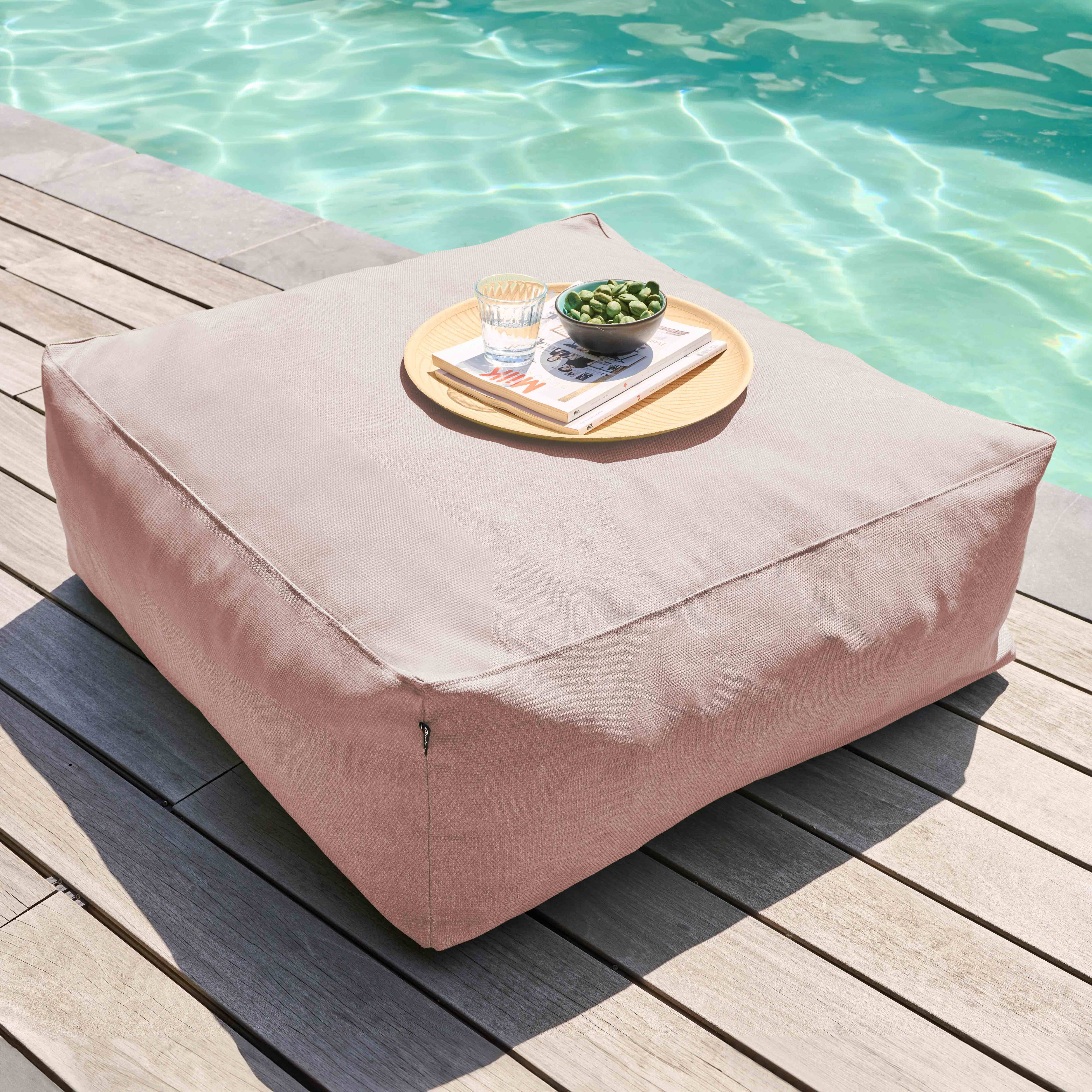 Ein beiges quadratisches Kissen auf Holzplanken neben einem Pool. Ein Tablett auf dem Kissen hält Snacks und ein Glas.