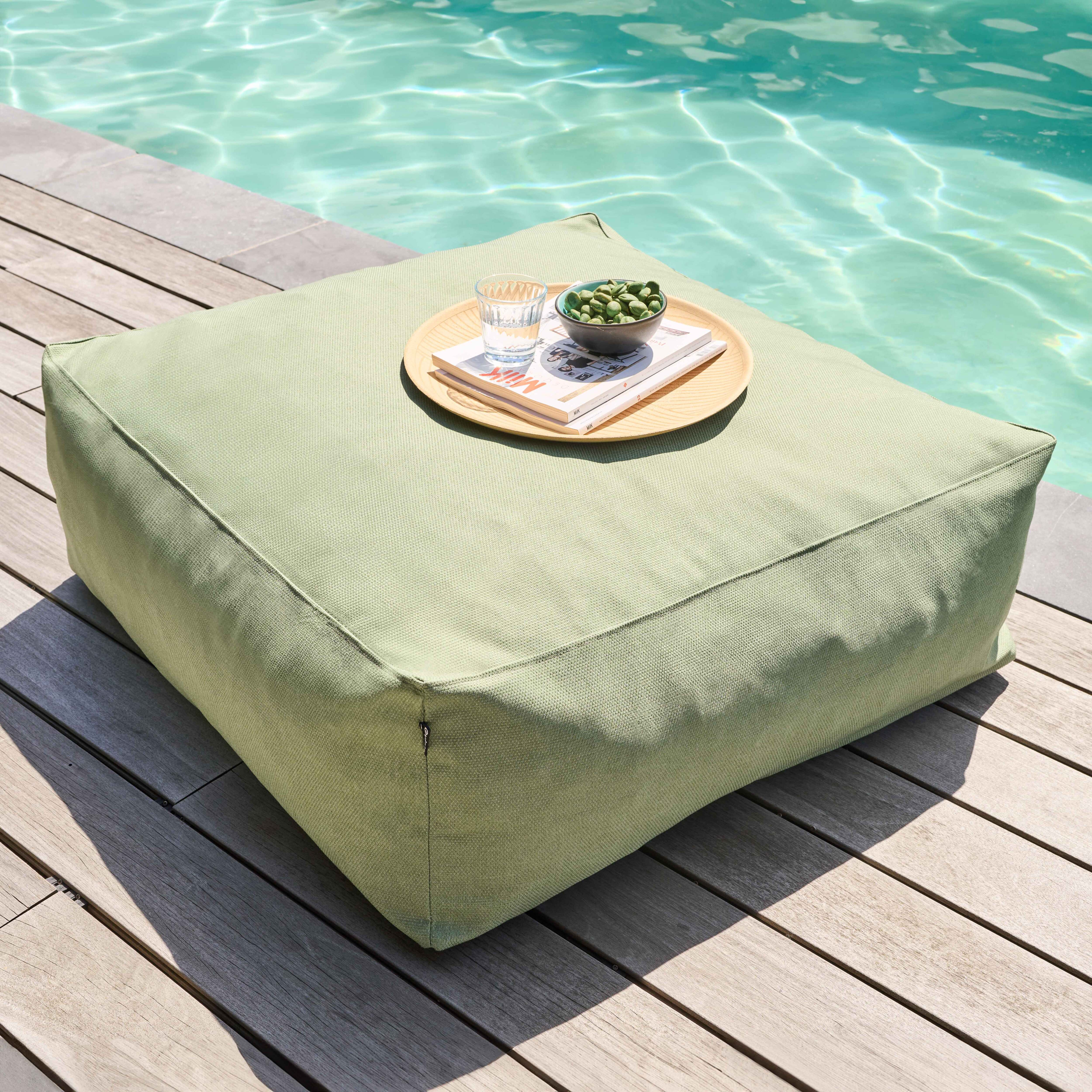 Ein grünes quadratisches Kissen mit einem Tablett mit Snacks steht auf Holzplanken neben einem Pool.