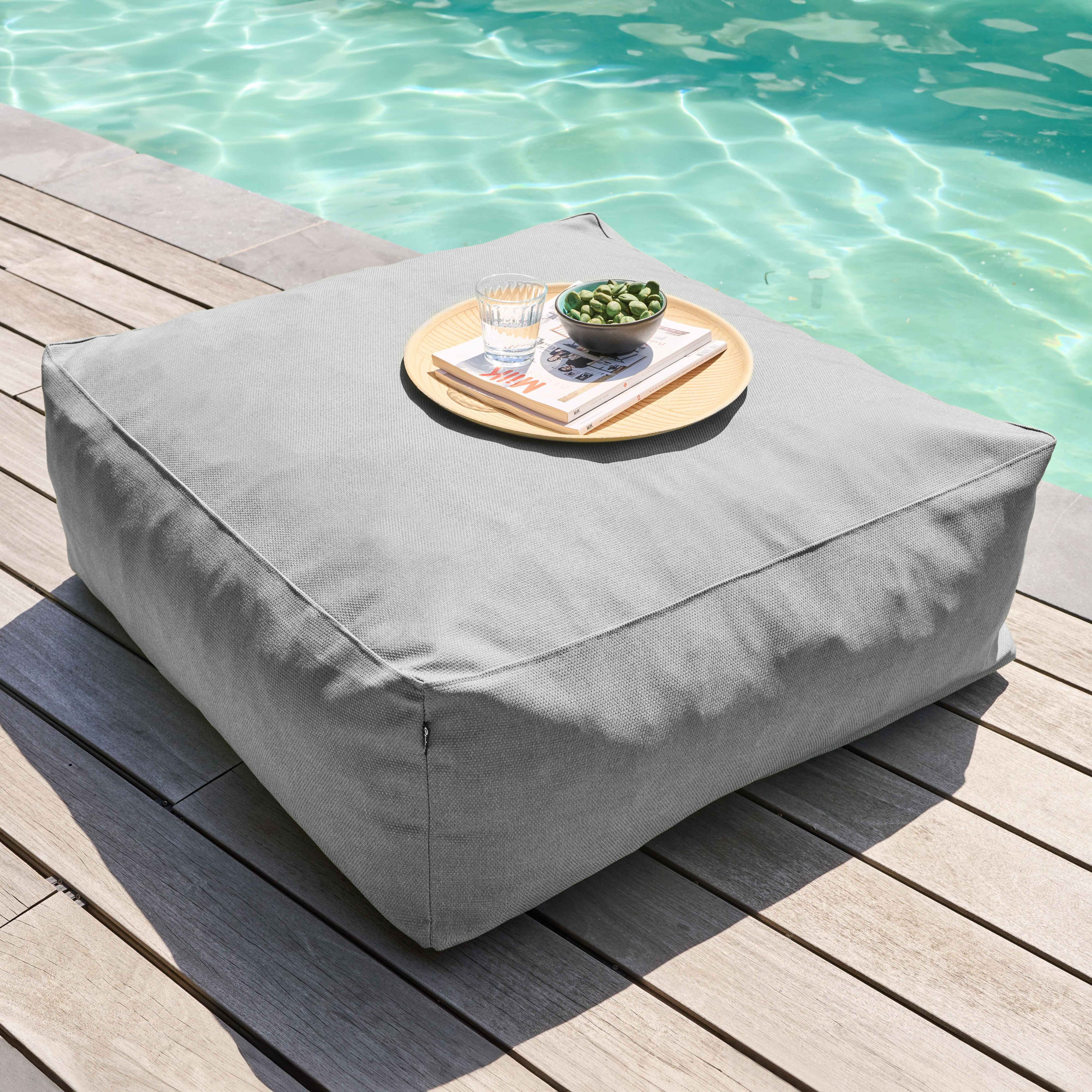 Graues quadratisches Outdoor-Kissen mit Gegenständen auf einem Tablett: Glas Wasser, Schüssel mit grünen Gegenständen und Bücher.