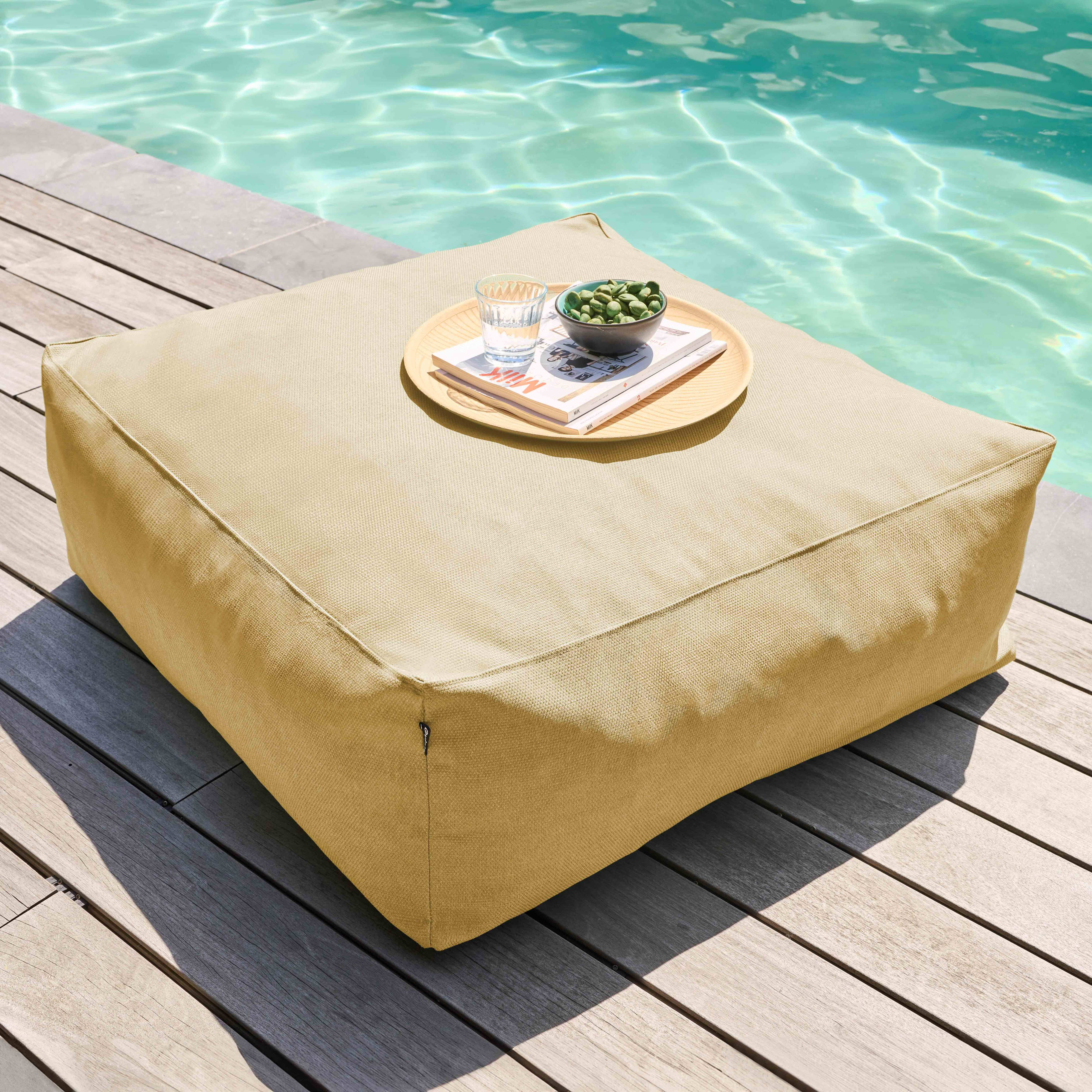 Ein beigefarbener quadratischer Hocker mit einem Tablett mit Snacks und einem Glas Wasser steht am Pool.