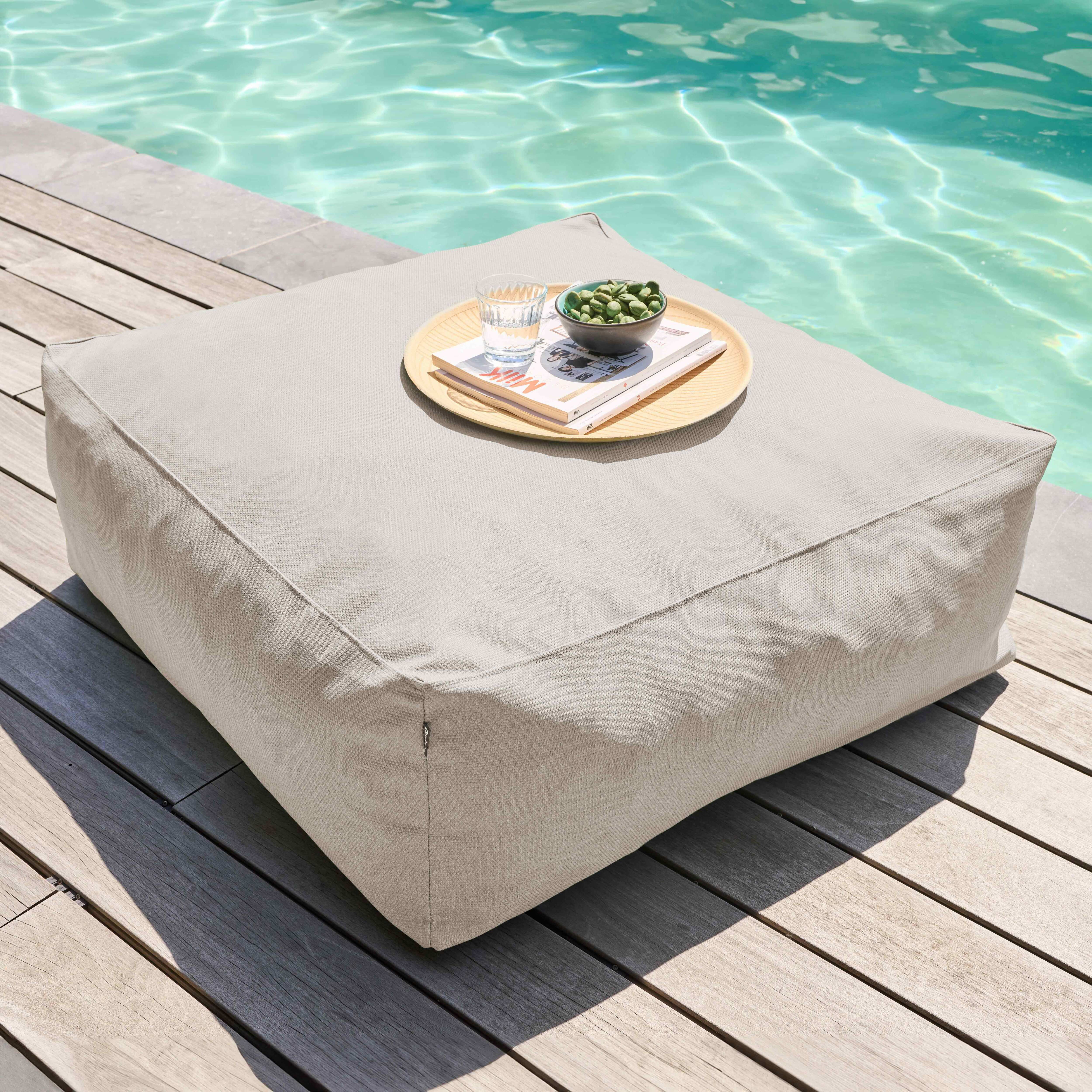 Ein beigefarbener quadratischer Ottoman mit einem Tablett mit Snacks und einem Wasserglas steht am Pool.