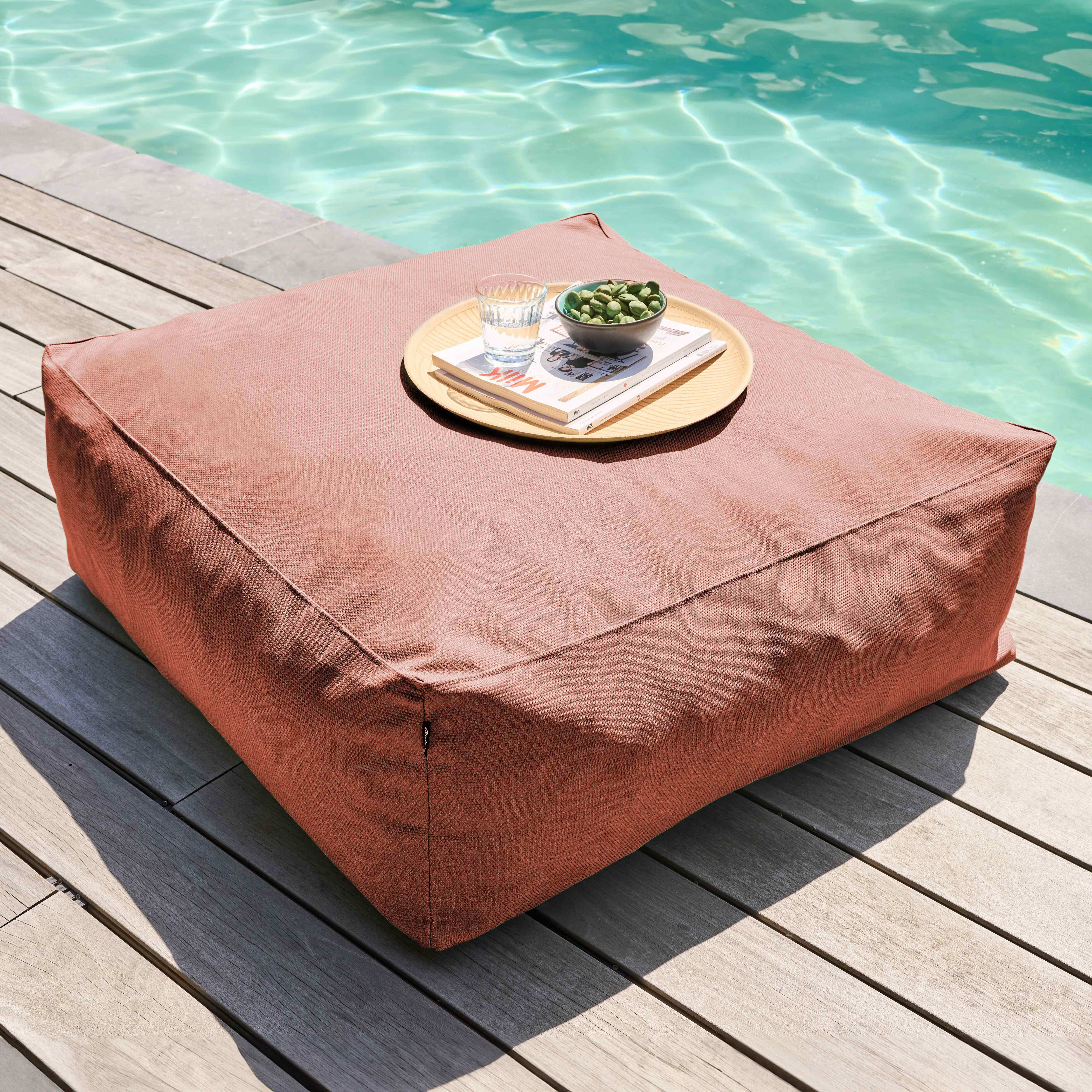 Ein großes, rosa Kissen auf Holzbrettern, mit einem Tablett mit Essen und einem Pool im Hintergrund.