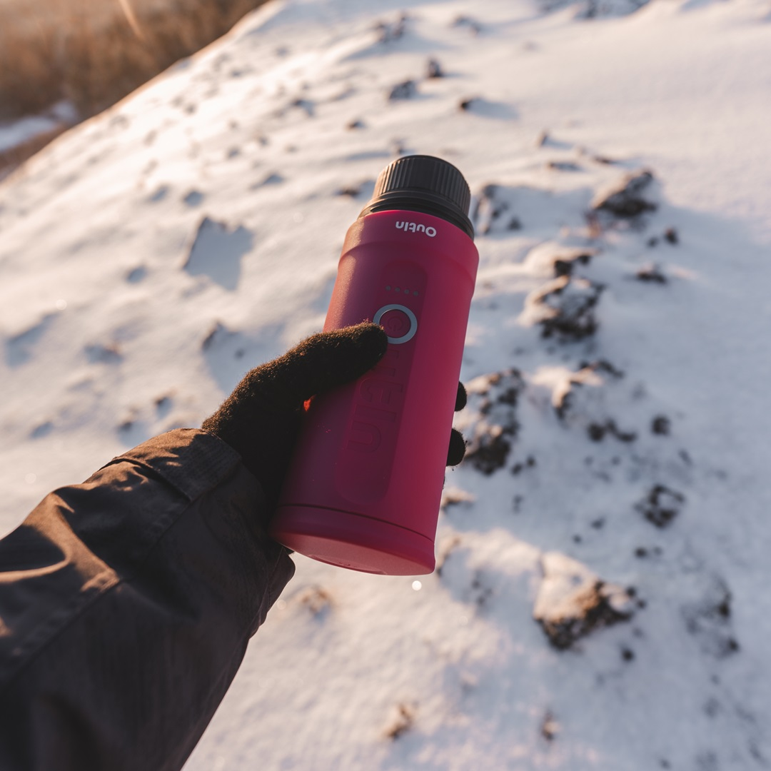 Ręka w czarnej rękawicy trzyma różową butelkę z wodą na tle zaśnieżonych gór.