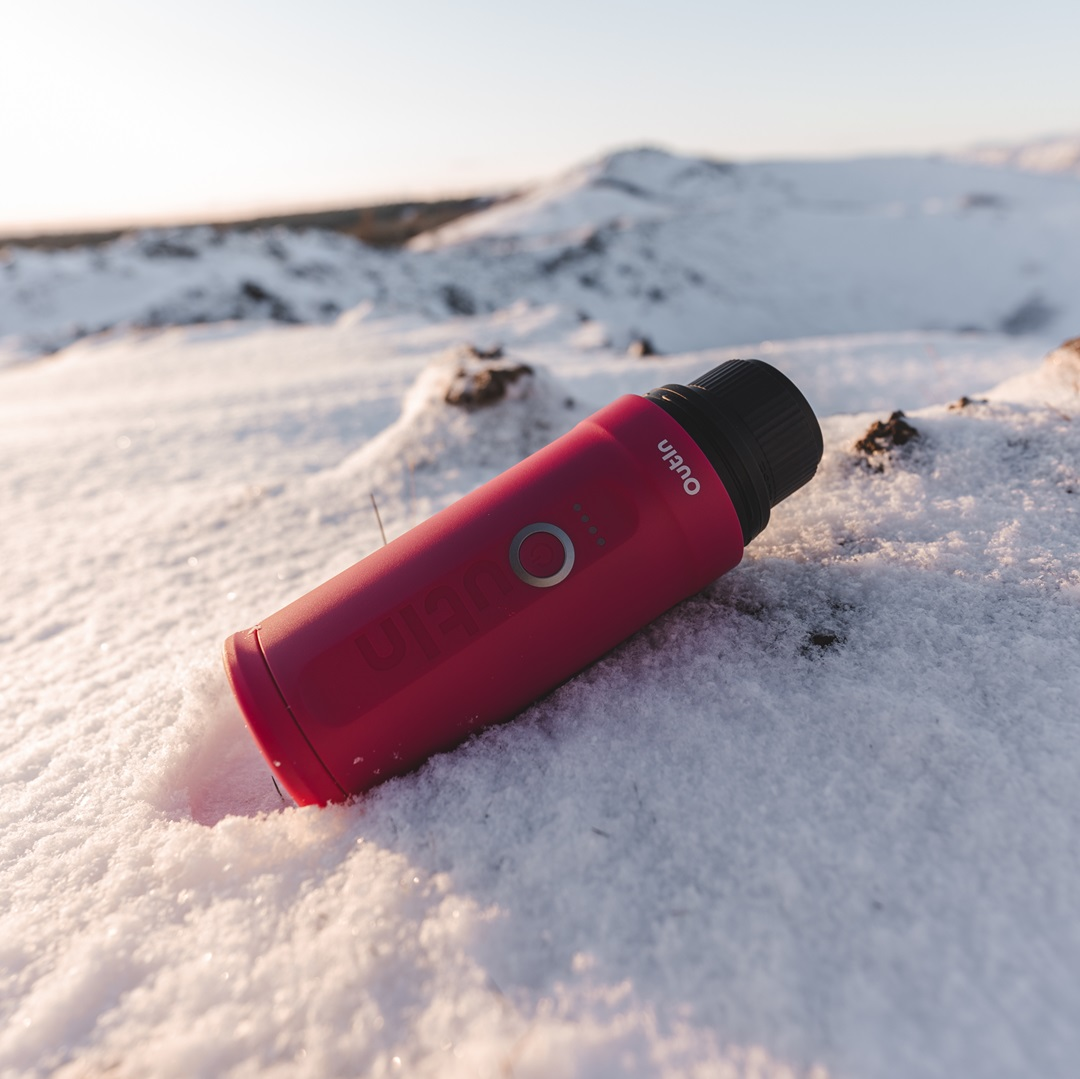 Czerwone urządzenie Outin spoczywa w śniegu. Czarna nasadka, przycisk z pierścieniem i wskaźniki zasilania są widoczne. Zaśnieżone góry w tle.