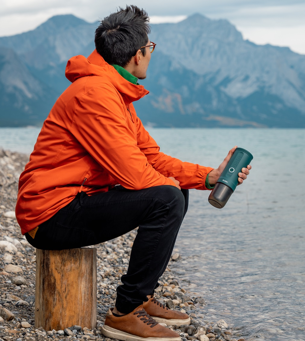 Mężczyzna w pomarańczowej kurtce siedzi na pniu nad jeziorem, trzymając butelkę z wodą, patrząc na góry w tle.