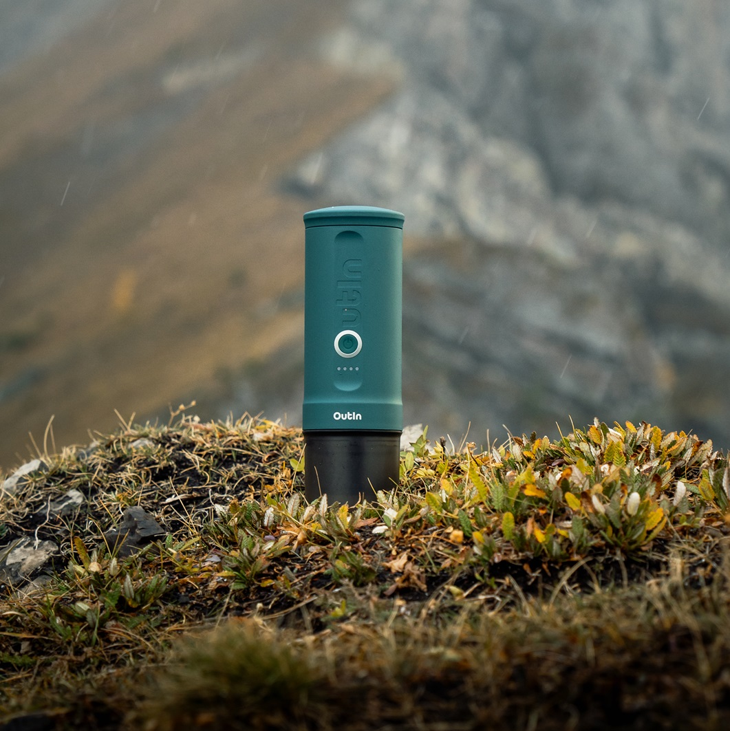 Turkusowe urządzenie Outin stoi na trawiastym wzgórzu, z górami w tle. Pada deszcz. Urządzenie jest cylindryczne, z białym przyciskiem.