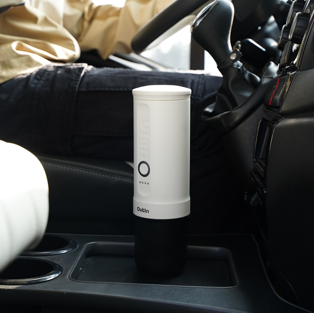 Een witte Outin-reismok staat in de console van een auto. Een persoon in een zwarte broek en een beige shirt zit op de bestuurdersstoel.