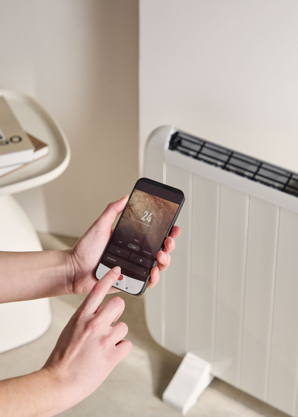 Una persona usa un teléfono para controlar un calentador. El teléfono muestra la configuración de temperatura. Calentador y mesa al fondo.