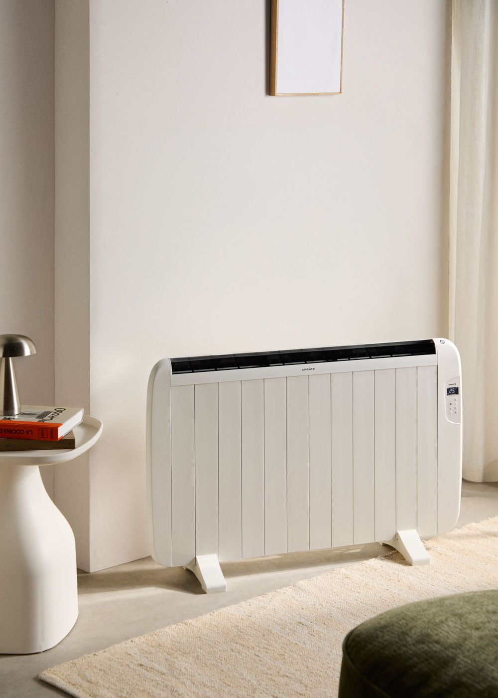 Un calefactor eléctrico blanco contra una pared blanca. A la izquierda, una mesita con libros y una lámpara.