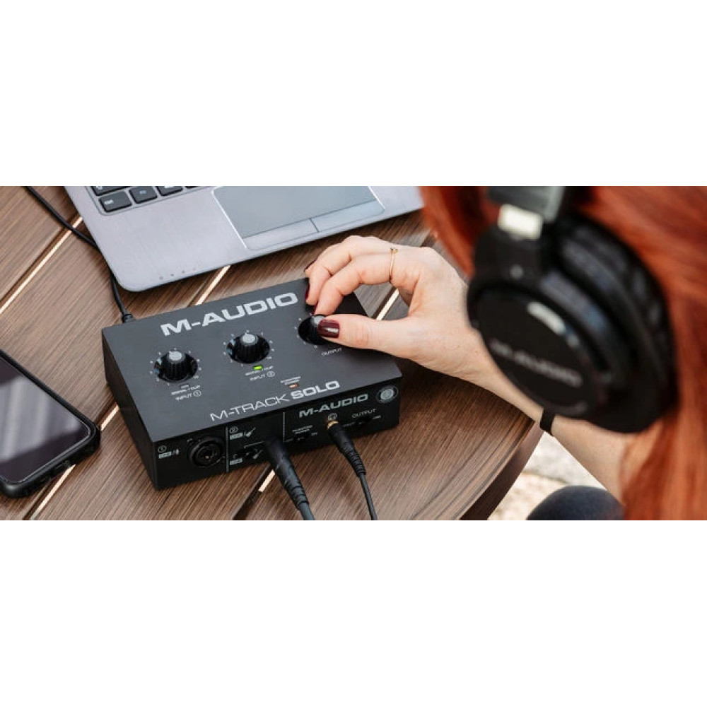 Osoba reguluje czarny interfejs audio M-Track Solo. Słuchawki po prawej. Laptop i telefon w pobliżu.