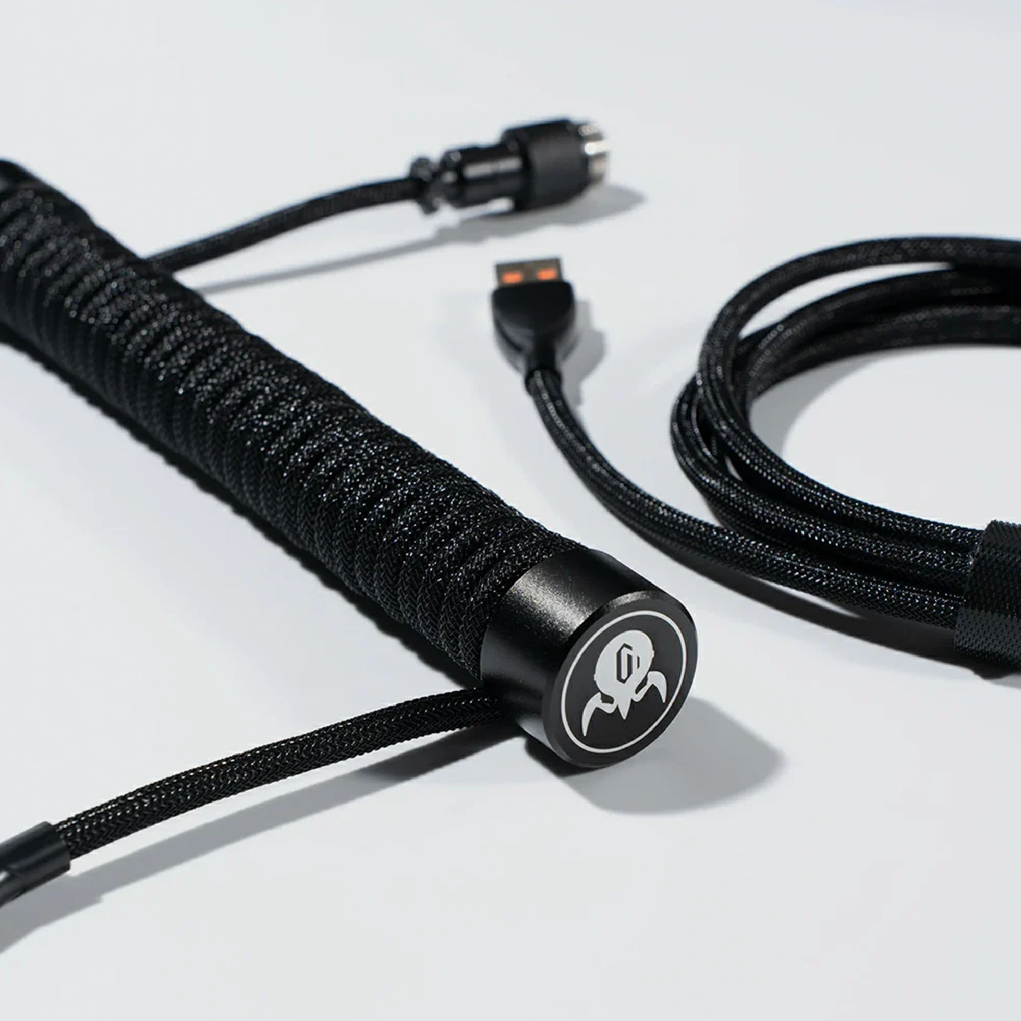 Beyaz yüzeyde USB ve konektör uçları olan siyah spiralli klavye kablosu. Bir ucunda logo var.