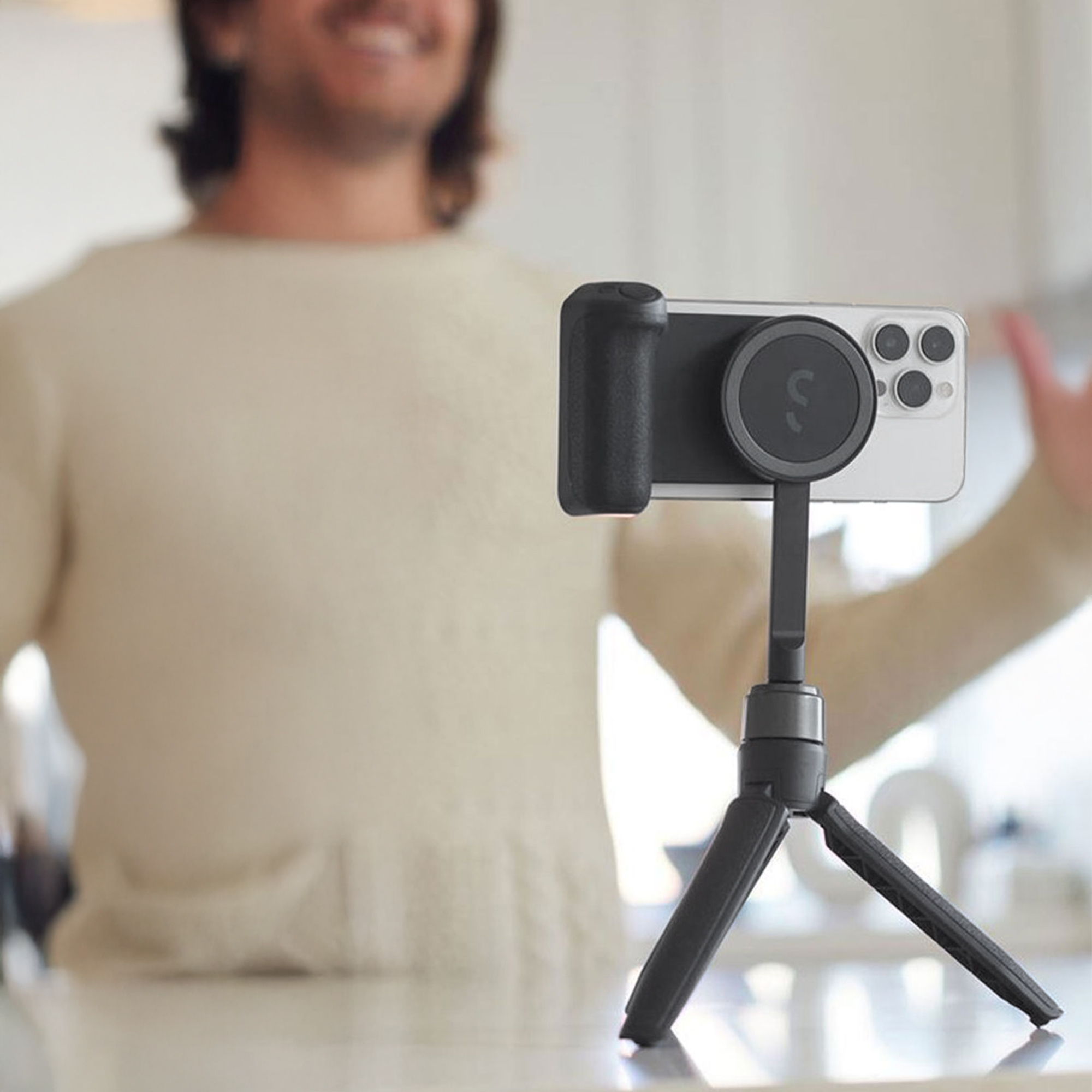 Tripod üzerindeki bir telefon net. Arka planda bir kişi bulanık, gülümsüyor ve jest yapıyor.