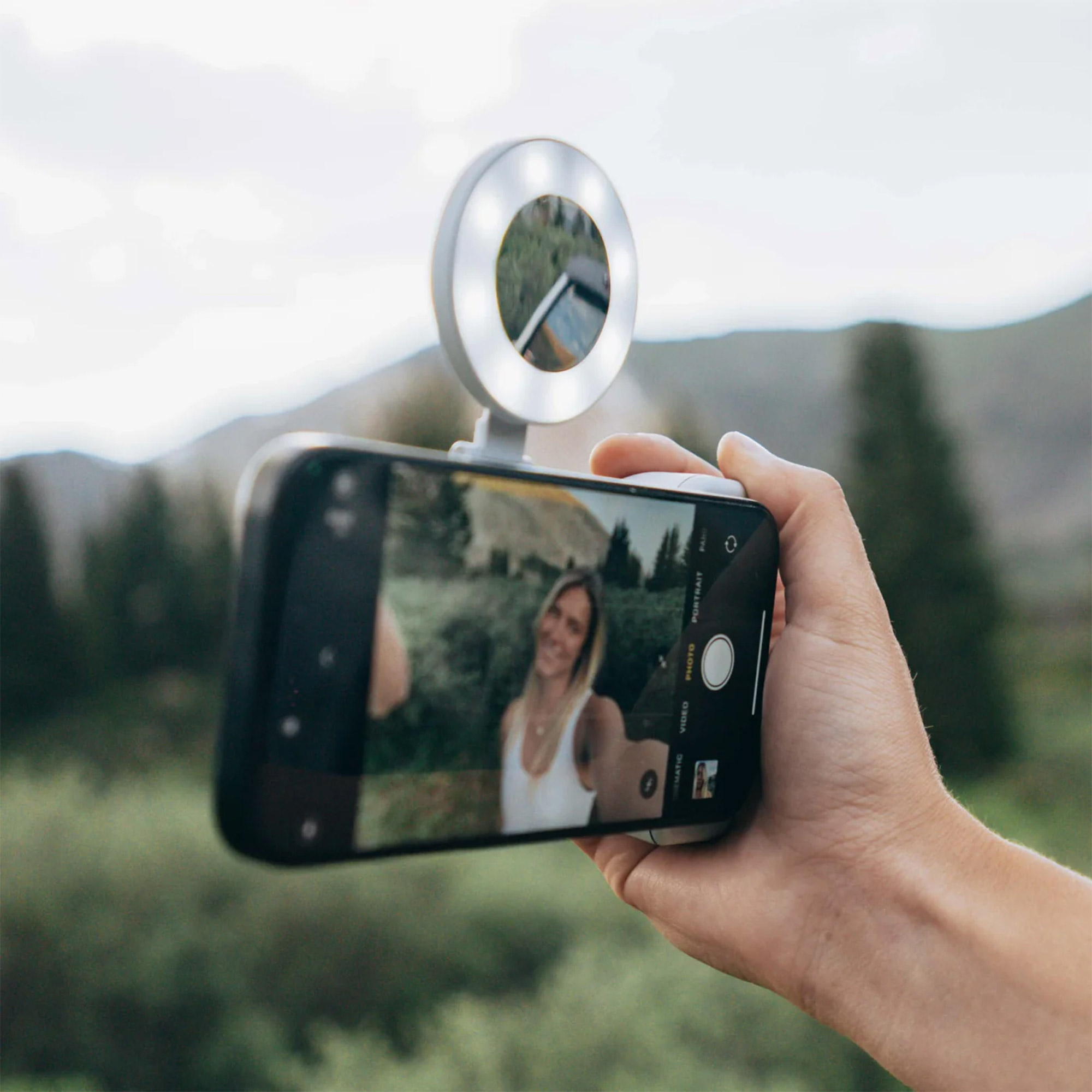 Bir el, selfie ve halka ışığı olan bir telefon tutuyor. Arka planda dağlar ve ağaçlar var.